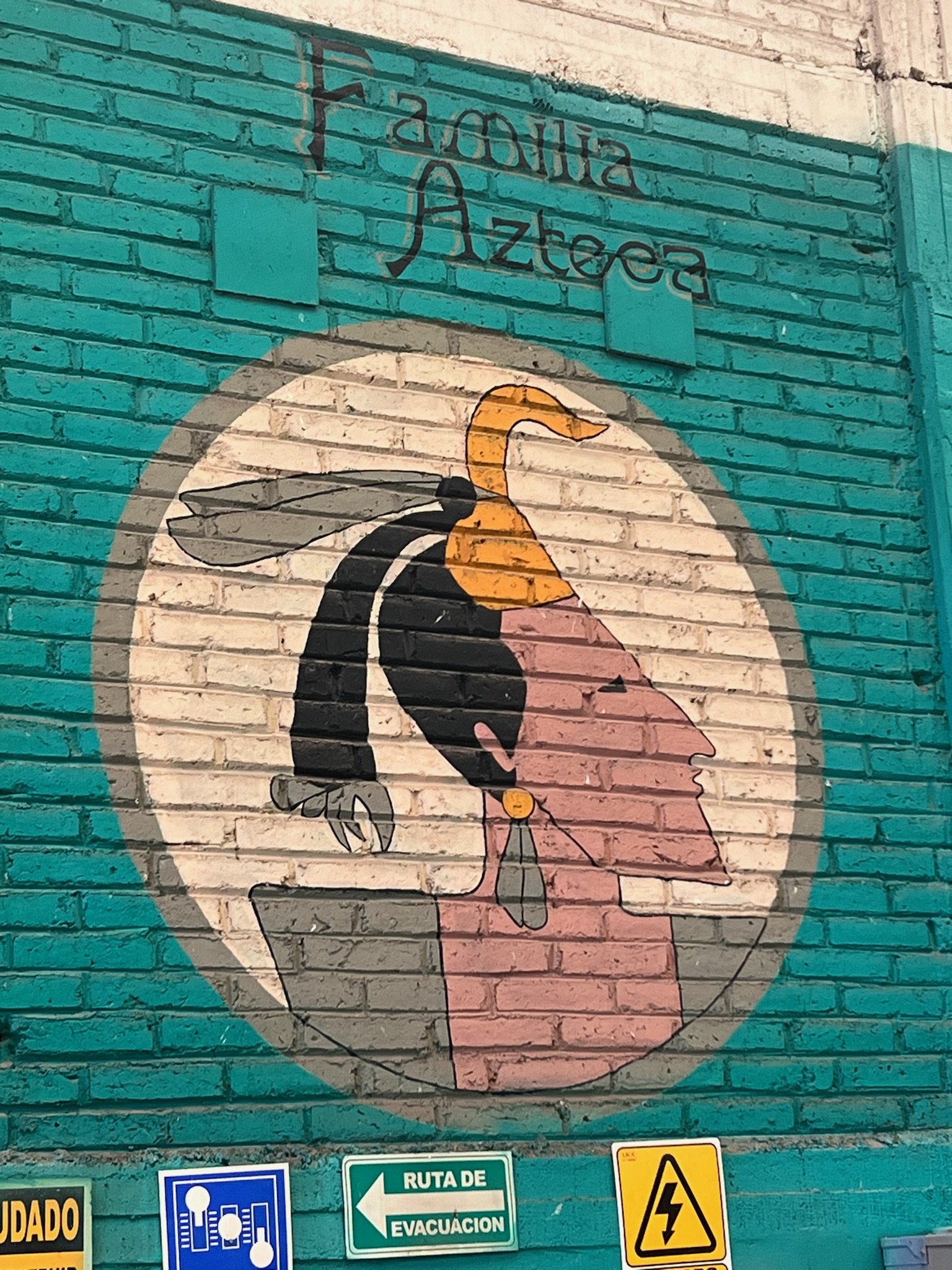 Aztec-themed mural on a brick wall with a profile of an Aztec warrior wearing a headdress. The text "Familia Azteca" is above, and various safety signs are below, including evacuation routes and caution signs.