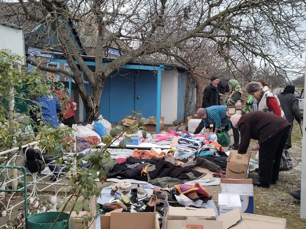 People engaging in a clothing swap or distribution event outdoors, with various clothes spread on tables and boxes nearby, likely during a cold season. A tree and a blue building are in the background.
