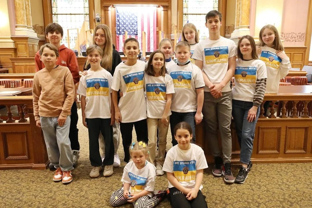 Group of children in a courtroom wearing "We Stand With Ukraine" shirts, with an American flag in the background.