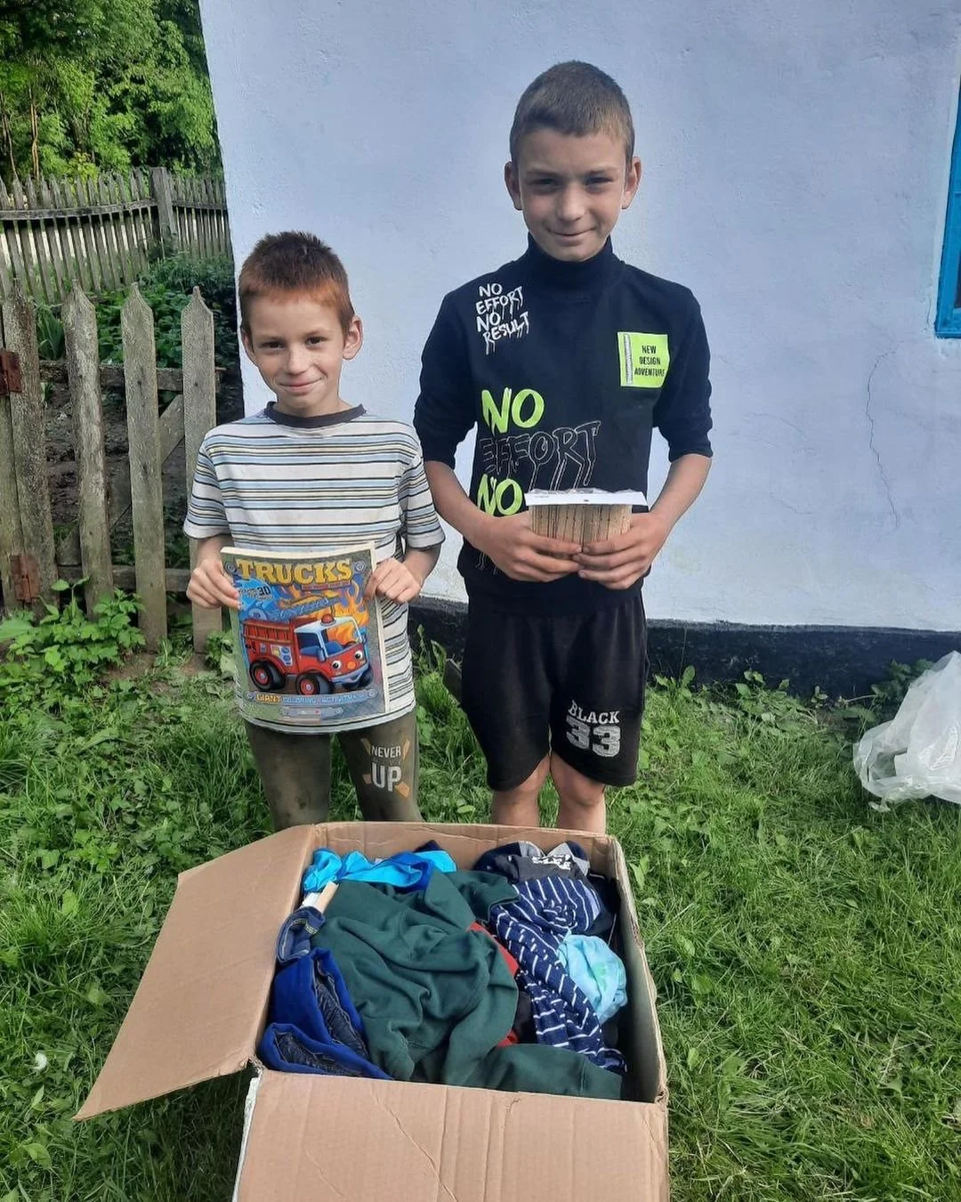 Two boys standing next to a cardboard box filled with assorted clothes. The boy on the left holds a trucks-themed activity book, and the boy on the right holds a stack of small boxes. They are outside near a wooden fence and a white building.