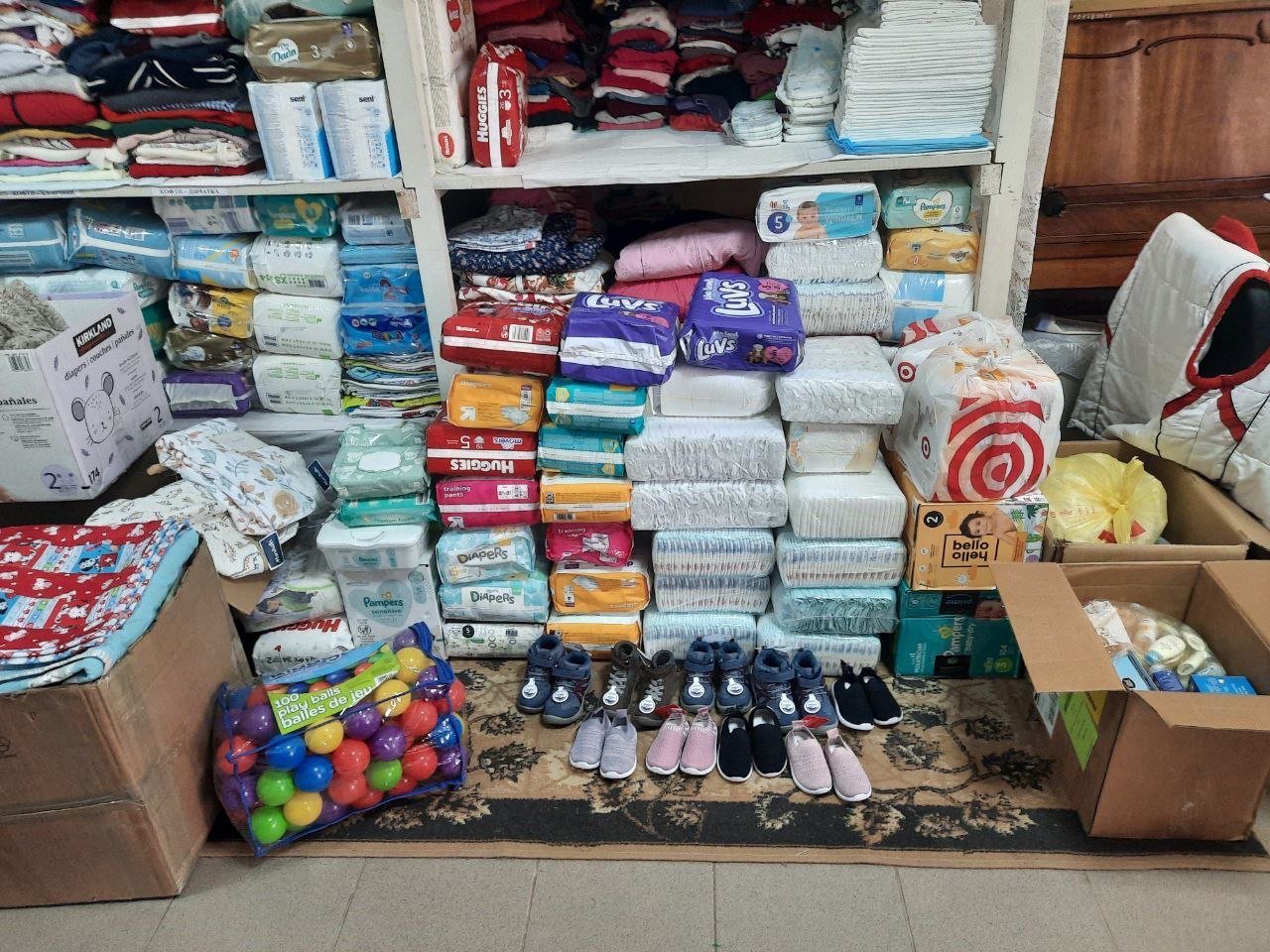 Stacks of baby diapers of various brands, children's shoes lined up, colorful toy balls in a bag, and assortment of folded baby clothes on shelves and in boxes.