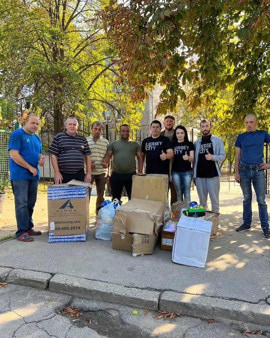 Group of people standing outdoors with boxes and bags on the ground, under a tree.