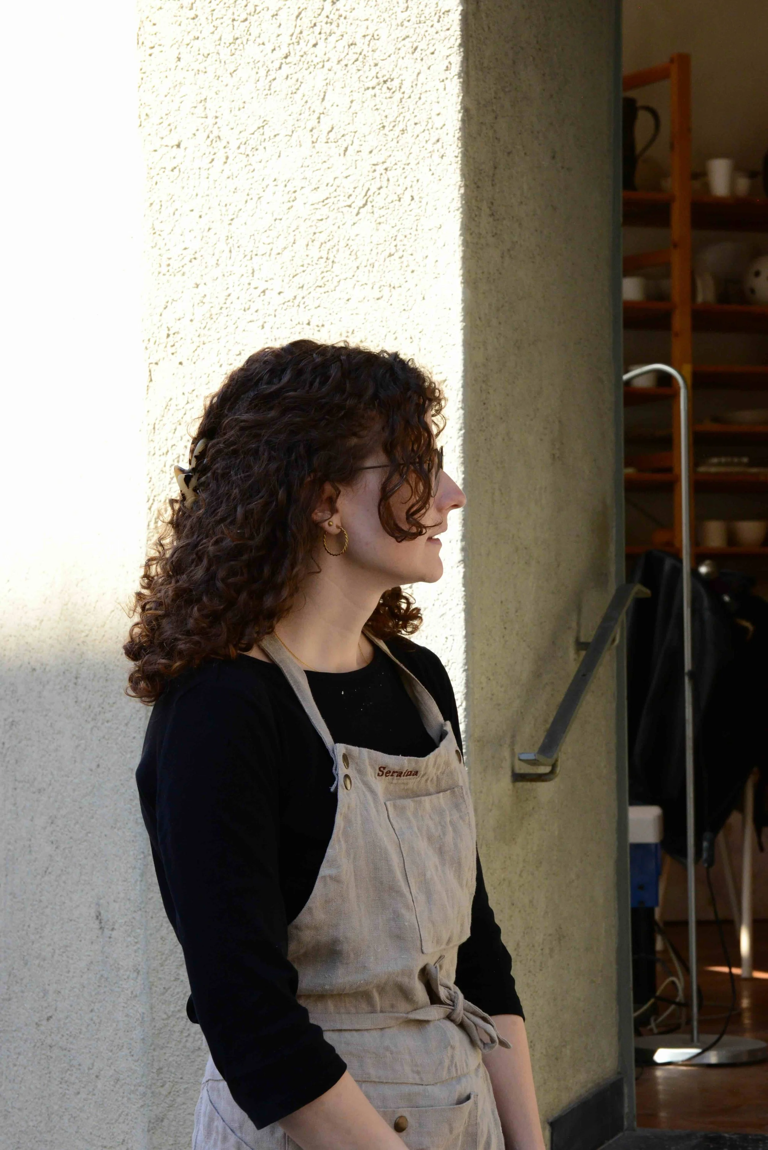 Frau mit lockigem braunem Haar, Brille und goldenen Ohrringen, trägt eine schwarze Oberbekleidung und eine beigen Schürze, steht seitlich vor einer hellen Wand in einem Raum mit Küchenutensilien im Hintergrund.