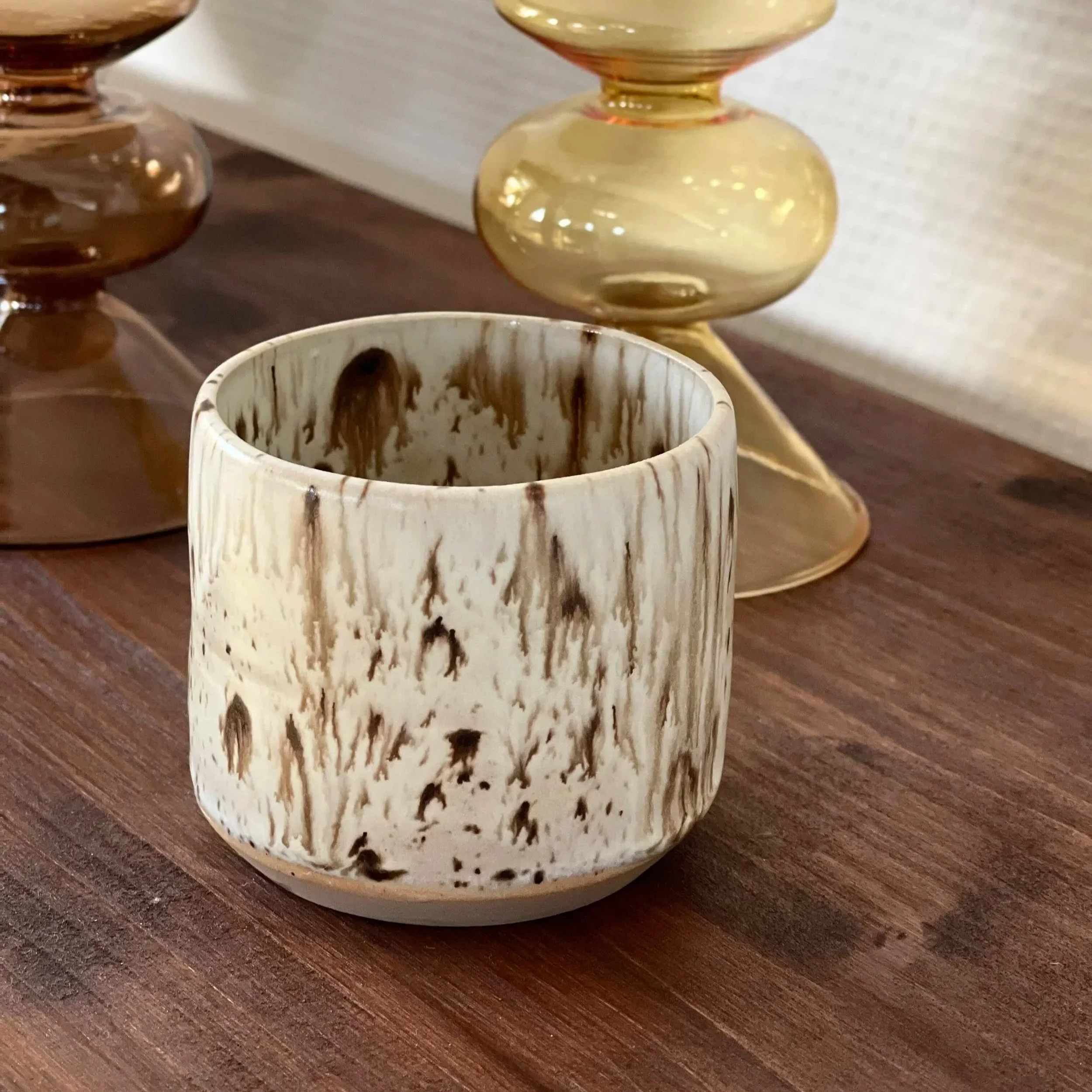 Keramikbecher mit braunen Muster, im Hintergrund zwei glasierte Kerzenständer in Cremé und Braun.