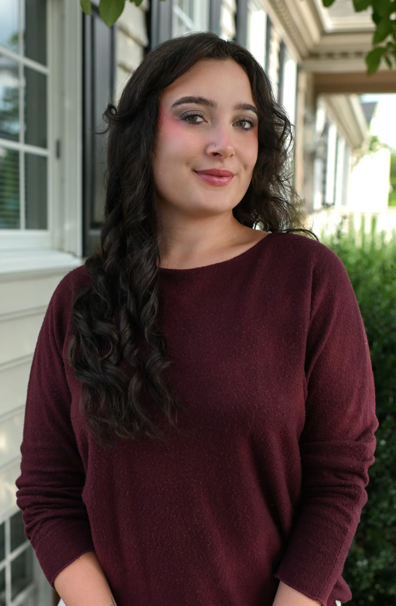 A young woman with dark curly hair, green eyes, and a nose piercing is standing outdoors near a house with large windows and greenery in the background. She is wearing a maroon long-sleeve top and smiling softly.