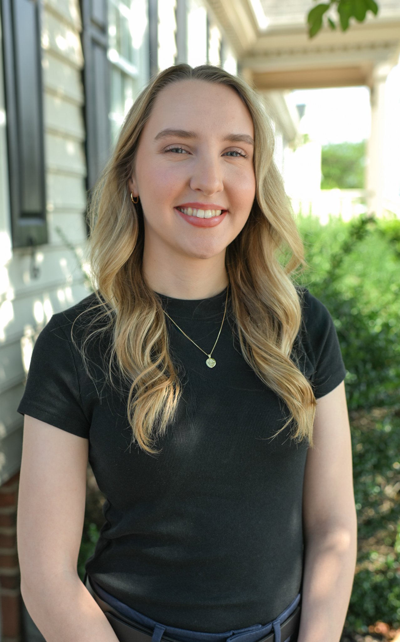 Young woman with blonde wavy hair smiling, wearing a black t-shirt, gold hoop earrings, and a gold necklace, standing outdoors with greenery and a porch in the background.