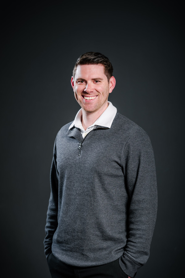 A smiling young man with dark hair, wearing a white collared shirt under a gray zippered sweater, standing against a dark gray background.