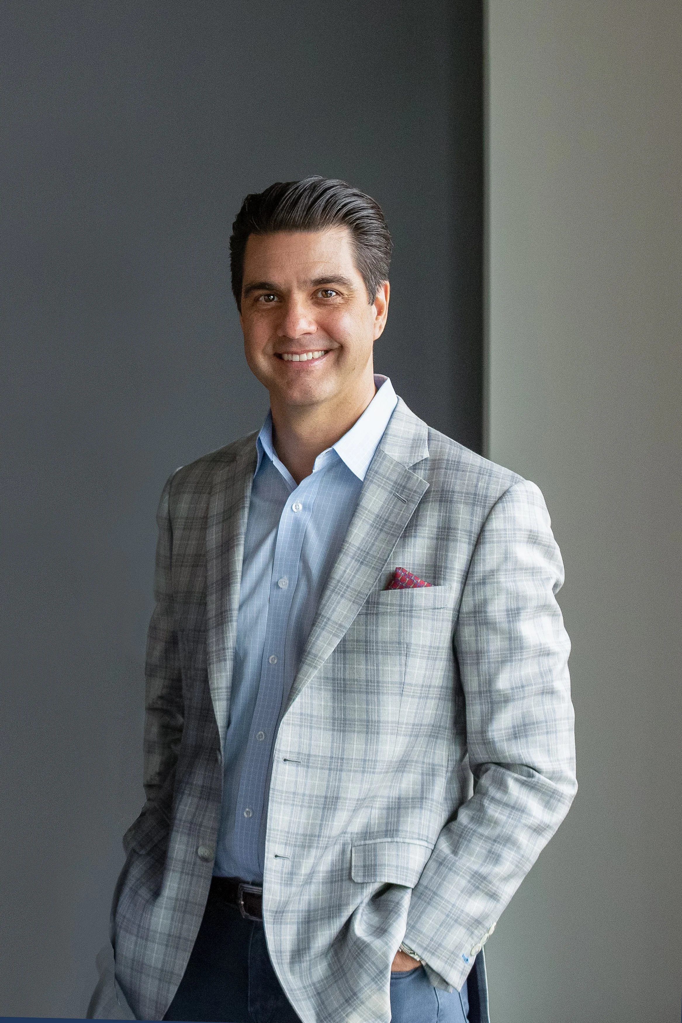 A smiling man in a light gray plaid suit jacket over a light blue dress shirt, standing in front of a dark and light gray wall.