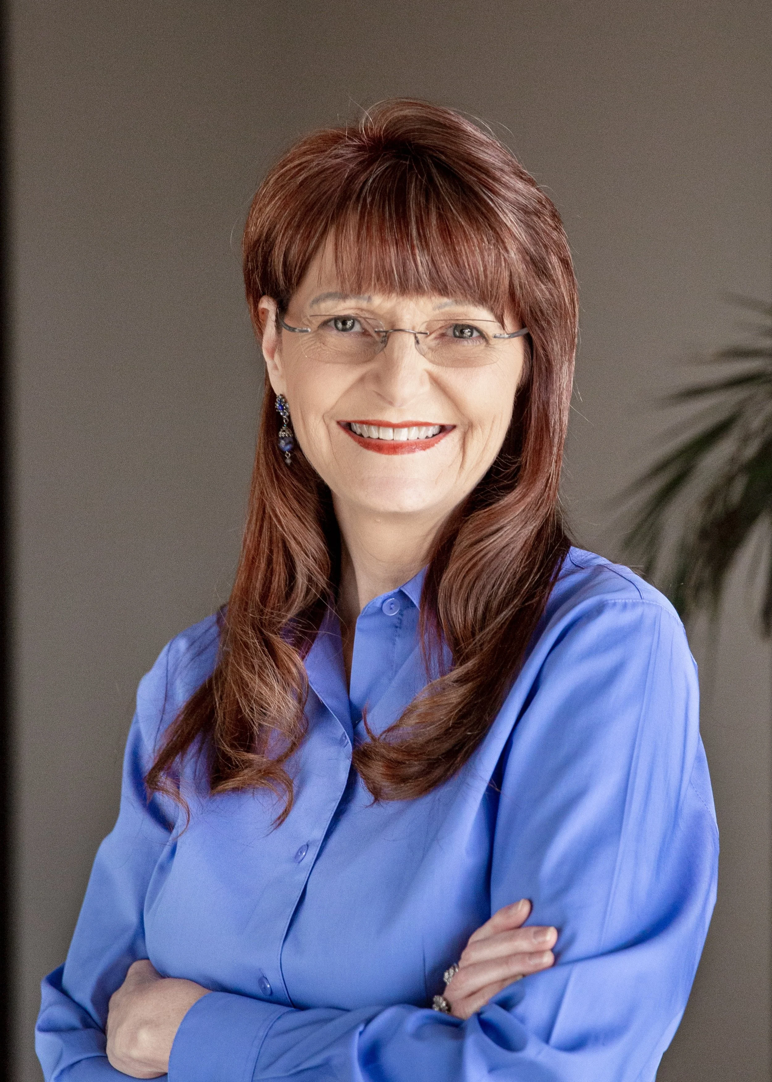 A smiling woman with red hair, glasses, and earrings, wearing a blue button-up shirt, standing with her arms crossed in front of a neutral background and a plant.