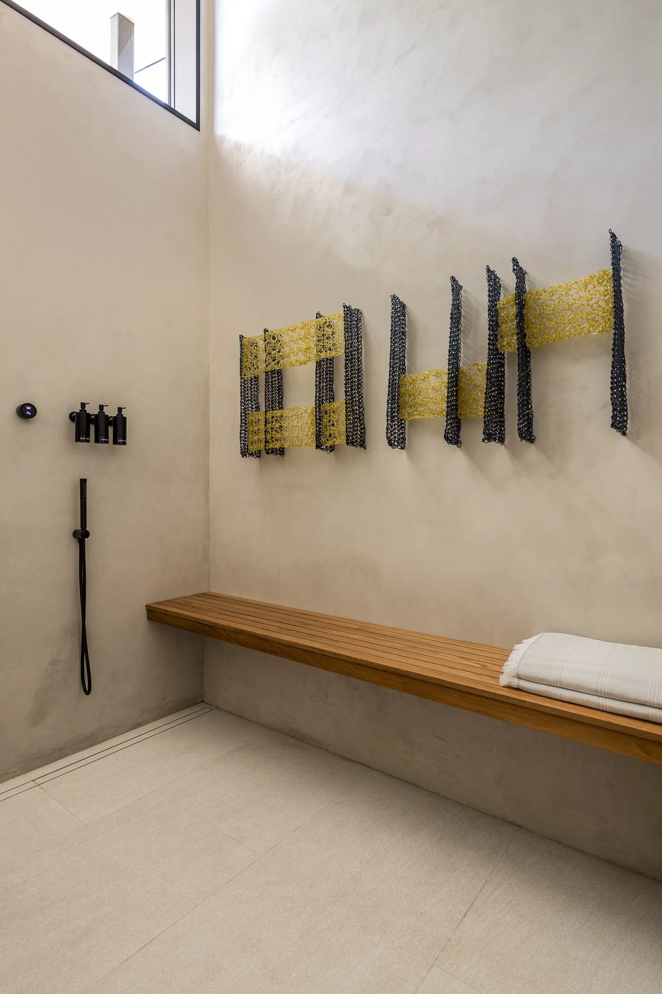 Modern, minimalist shower room with beige textured walls, a wooden bench, black wall-mounted shower fixtures, and decorative black and gold lattice panels on the wall, with natural light coming in from a nearby window.