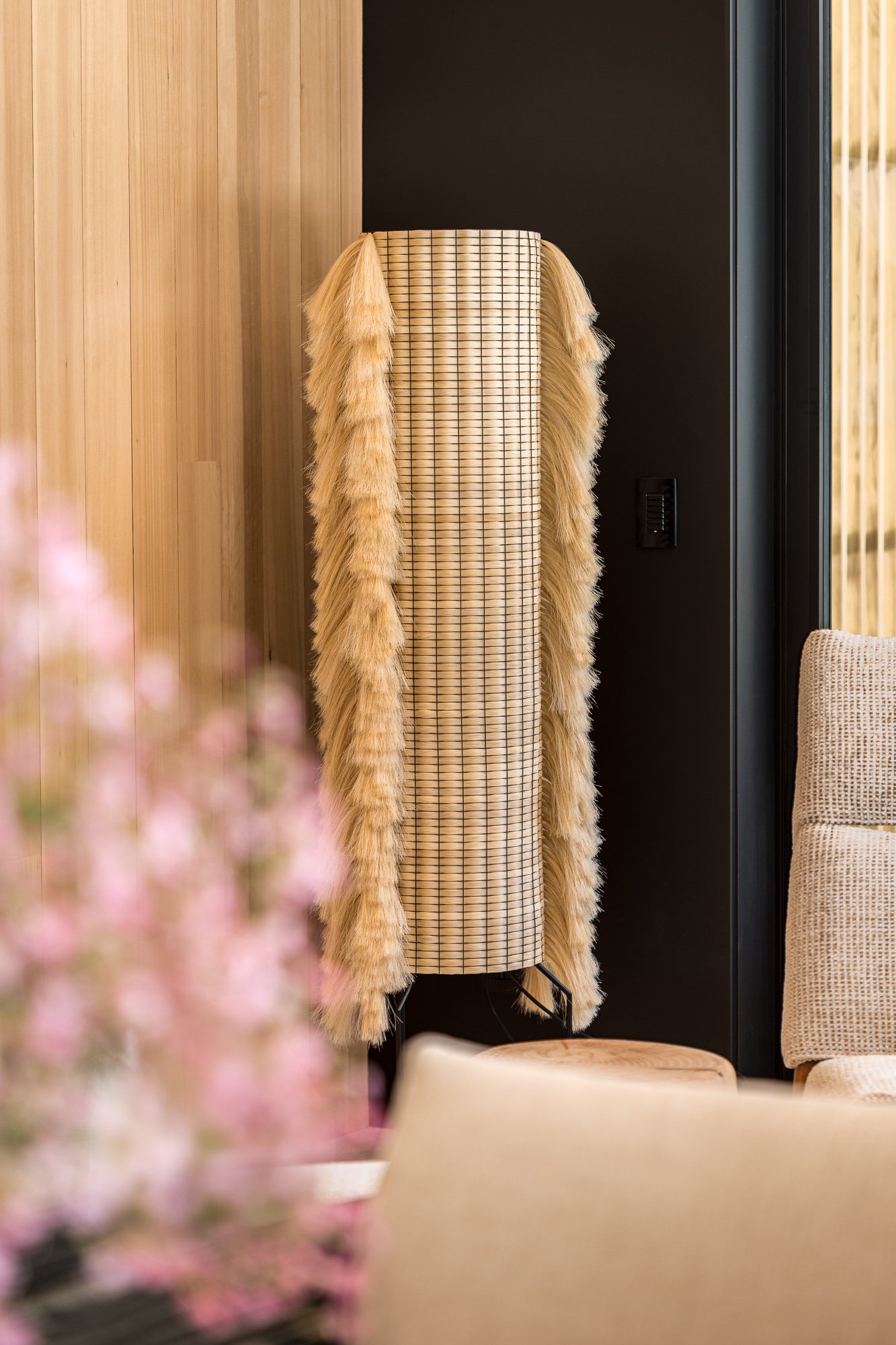 A coat rack with a beige and black checkered coat and a long fur stole, situated near a black wall and a glass door, with a beige chair and a wooden table in the foreground.