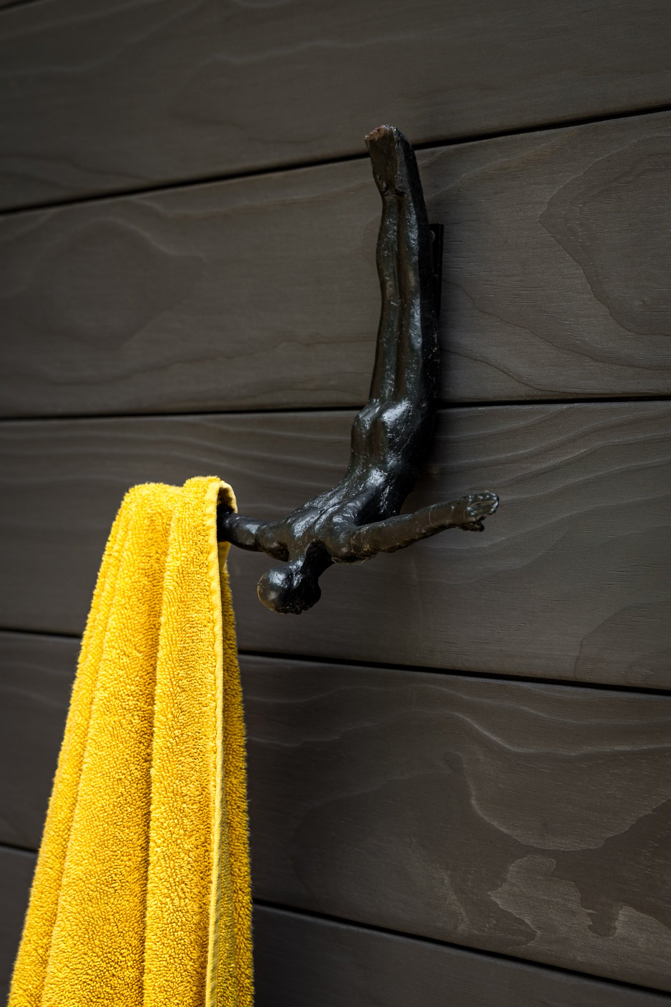 Bronze sculpture of a dog stretching or playing, mounted on a wall with dark horizontal wooden panels. A yellow towel hangs from the sculpture.