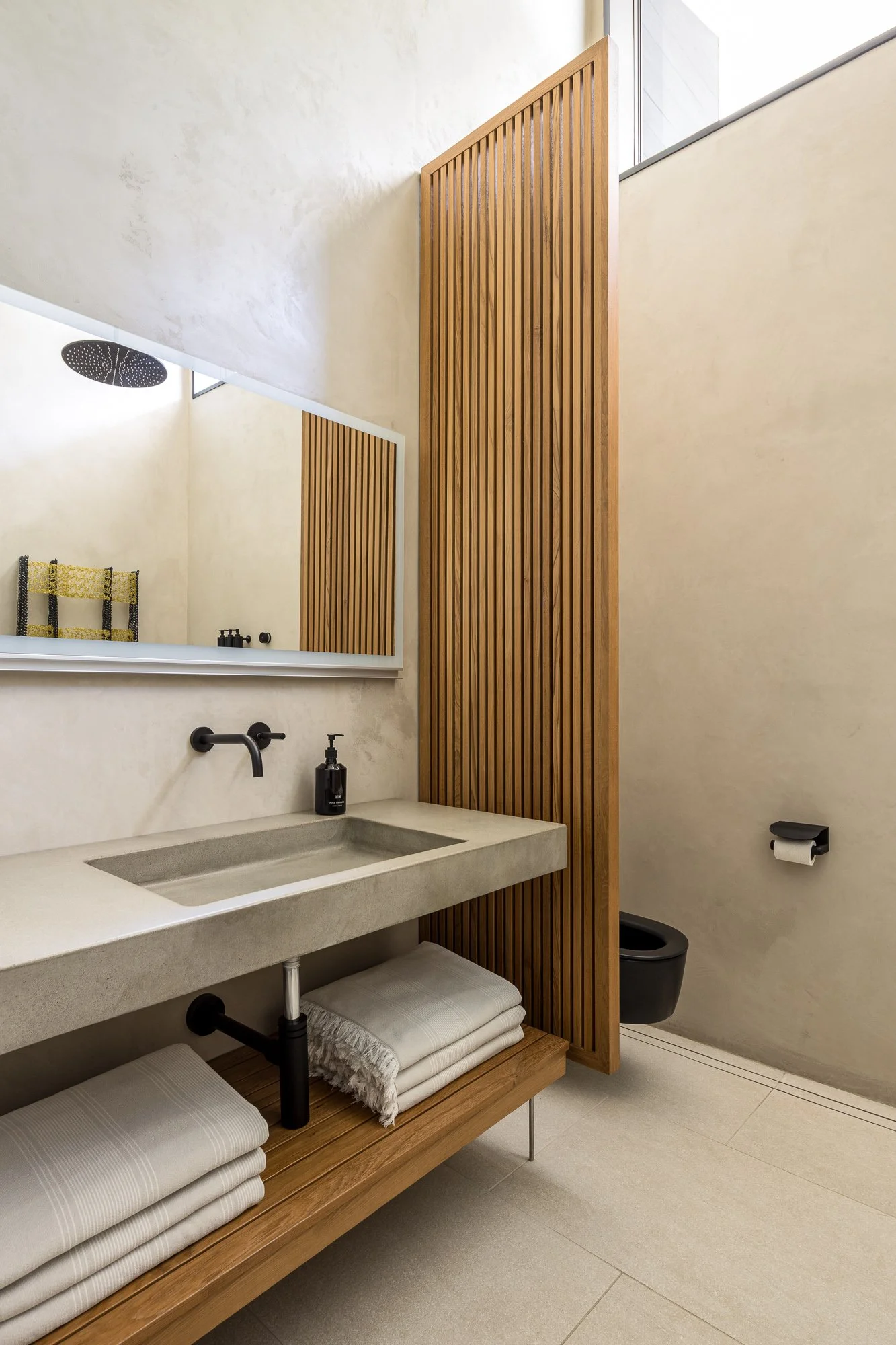 A modern bathroom with a beige concrete sink, black faucet, large mirror, wooden slatted divider, towel shelf underneath, and a black toilet with a roll of toilet paper.