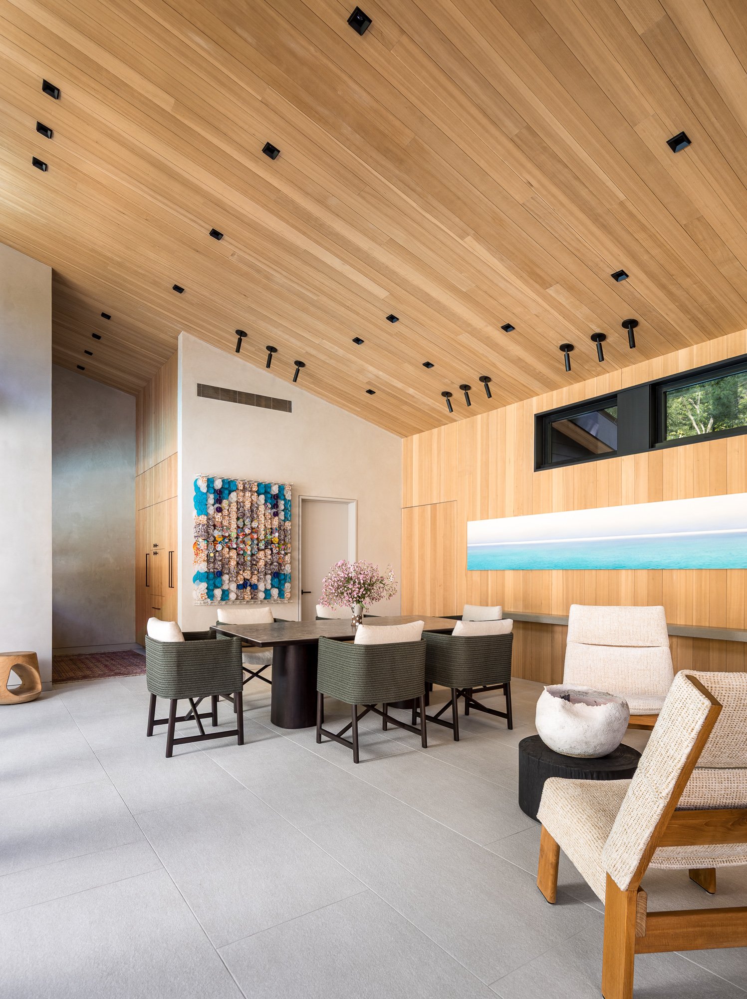 Interior of a modern room featuring wooden ceiling panels with black recessed lighting, a large window, a table with chairs, and contemporary furniture including a beige armchair and a decorative sculpture.
