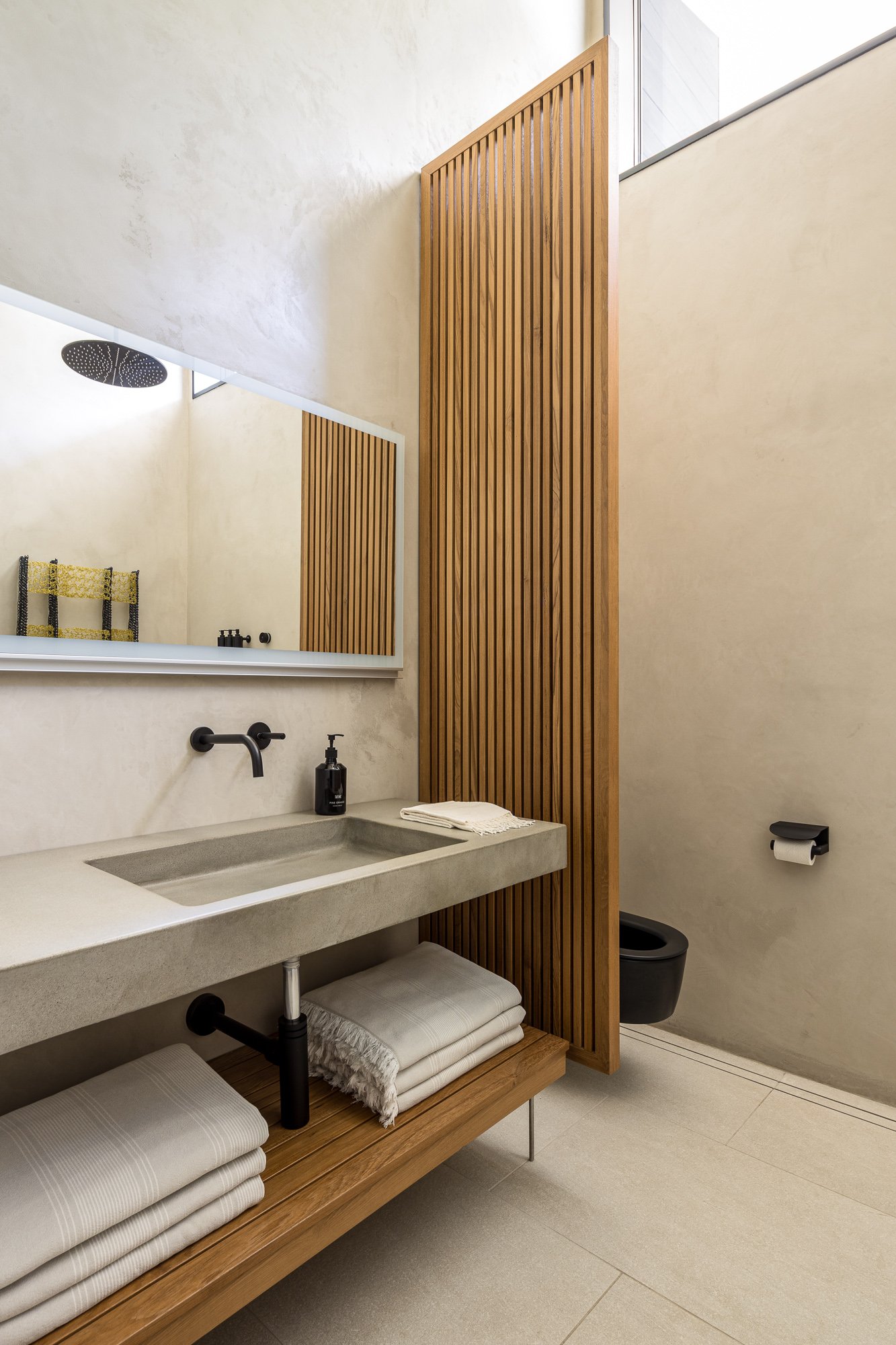 Modern bathroom with a concrete sink, wooden slatted partition, black toilet, black faucet, large mirror, and beige walls and tile flooring.