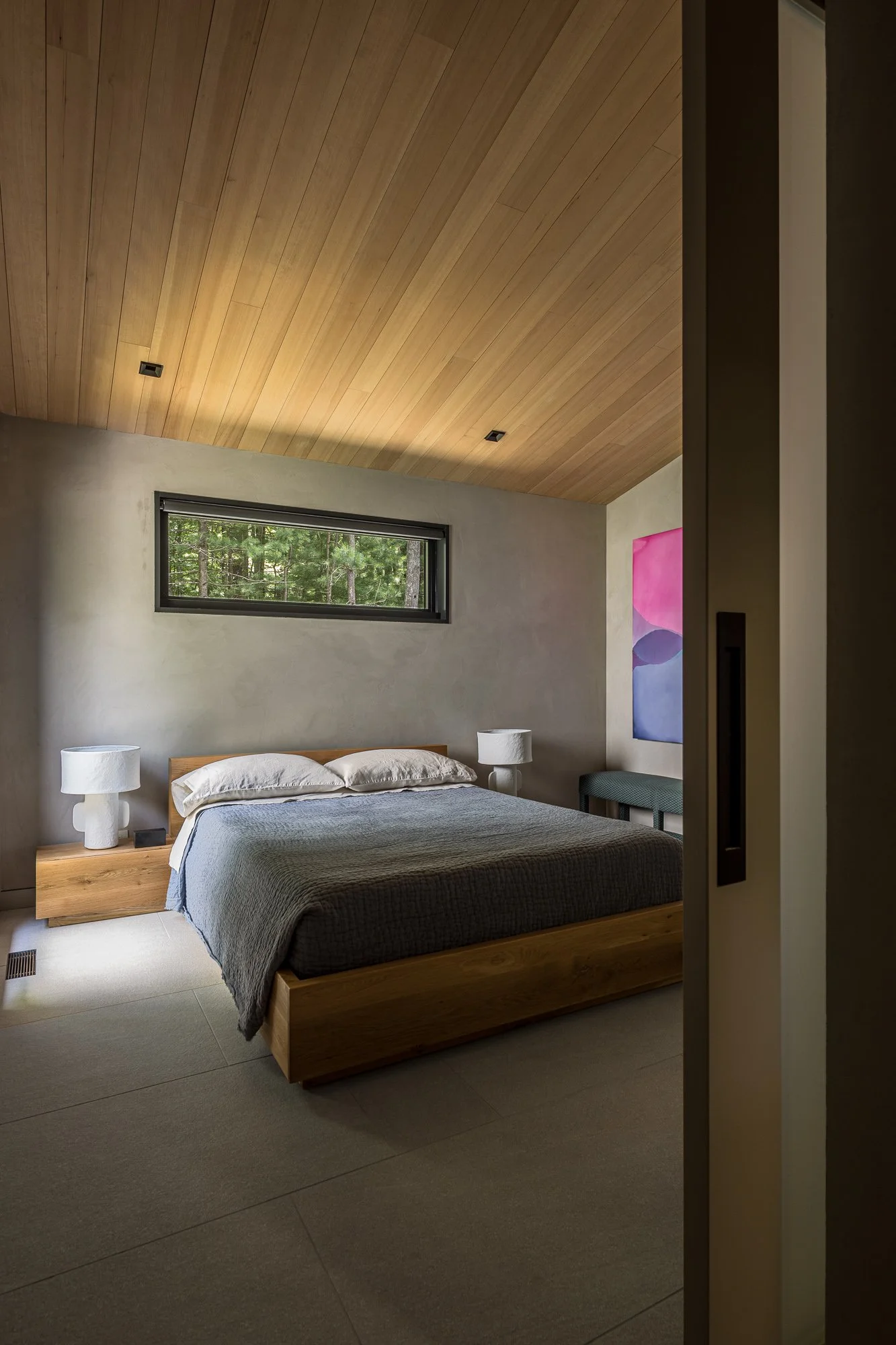 A modern bedroom with a wooden ceiling, a horizontal window showing trees outside, a bed with a wooden frame and grey bedding, two white bedside lamps, and a colorful abstract painting on the wall.