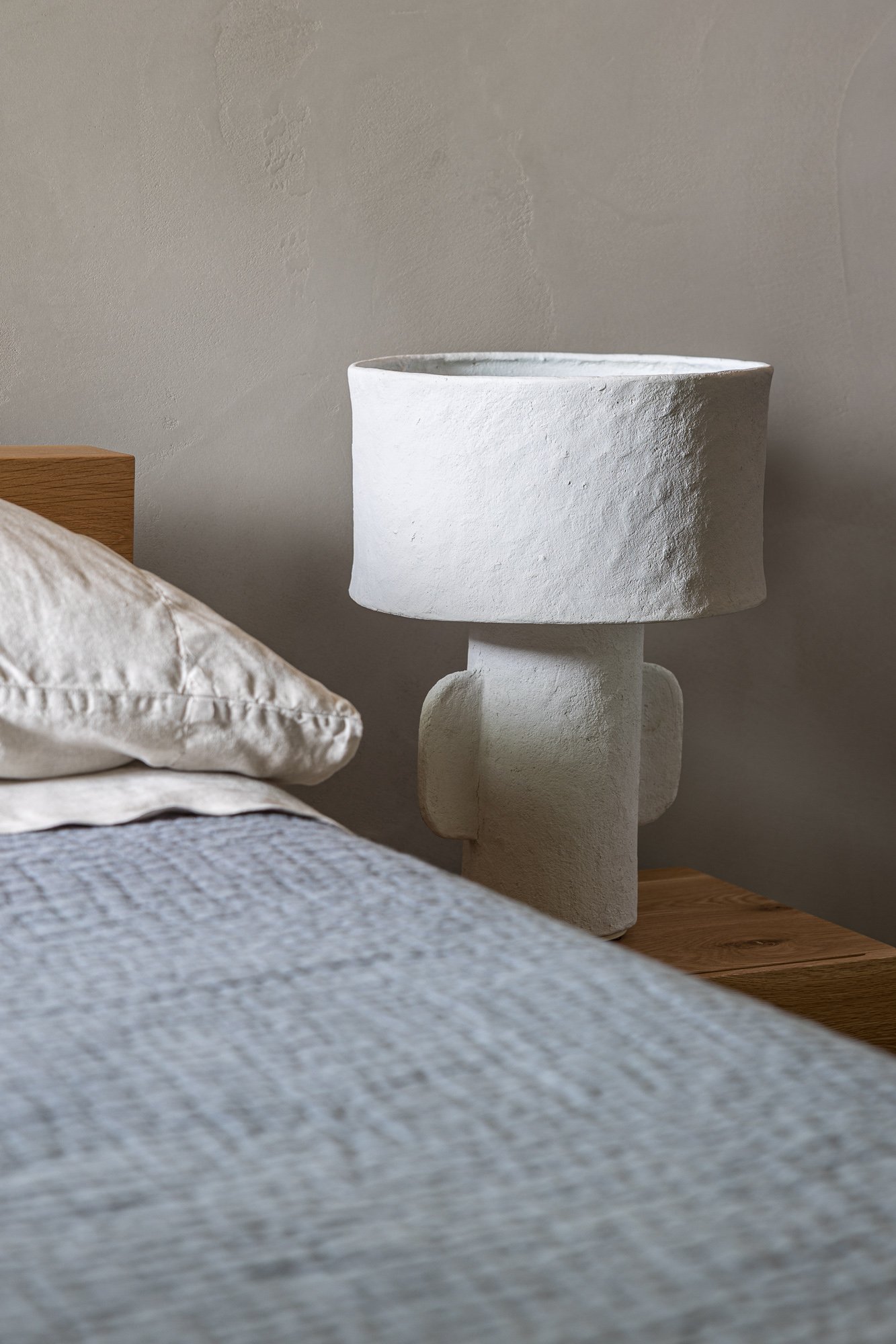 Close-up of a textured white bedside table with a large white pillow and a knitted blanket on a bed in the foreground, and a beige wall in the background.