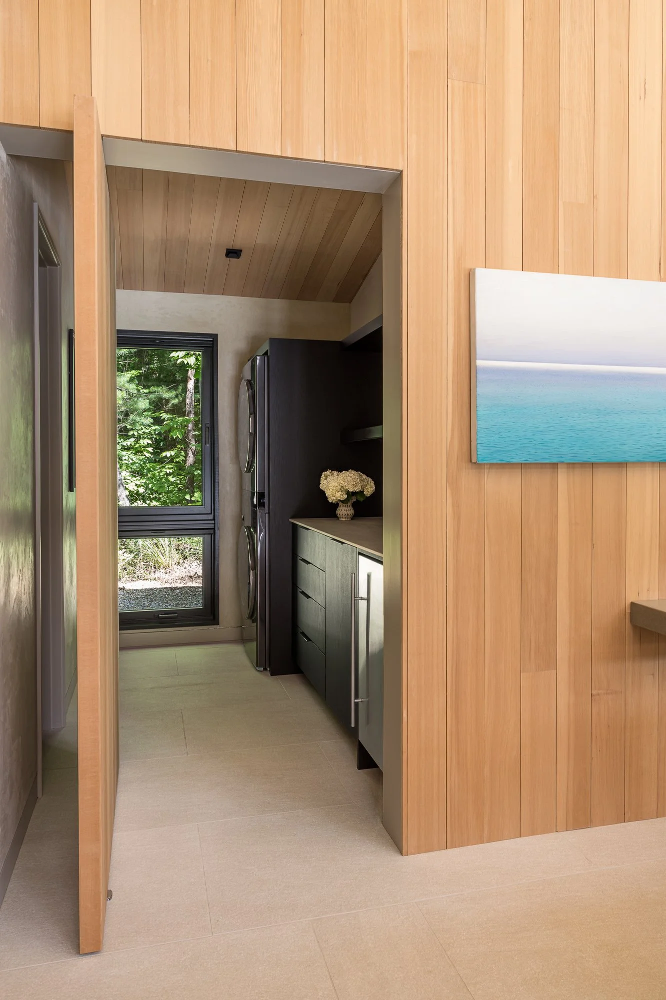A view of a laundry room with washer and dryer units, a window showing greenery outside, and a decorative flower arrangement on a cabinet.