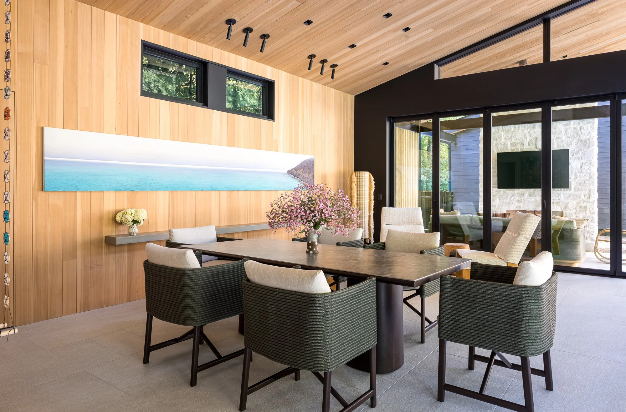 Modern dining room with a large wooden table surrounded by eight chairs, pink flowers in a vase, and a large landscape painting on a wooden wall, with sliding glass doors leading to an outdoor area.