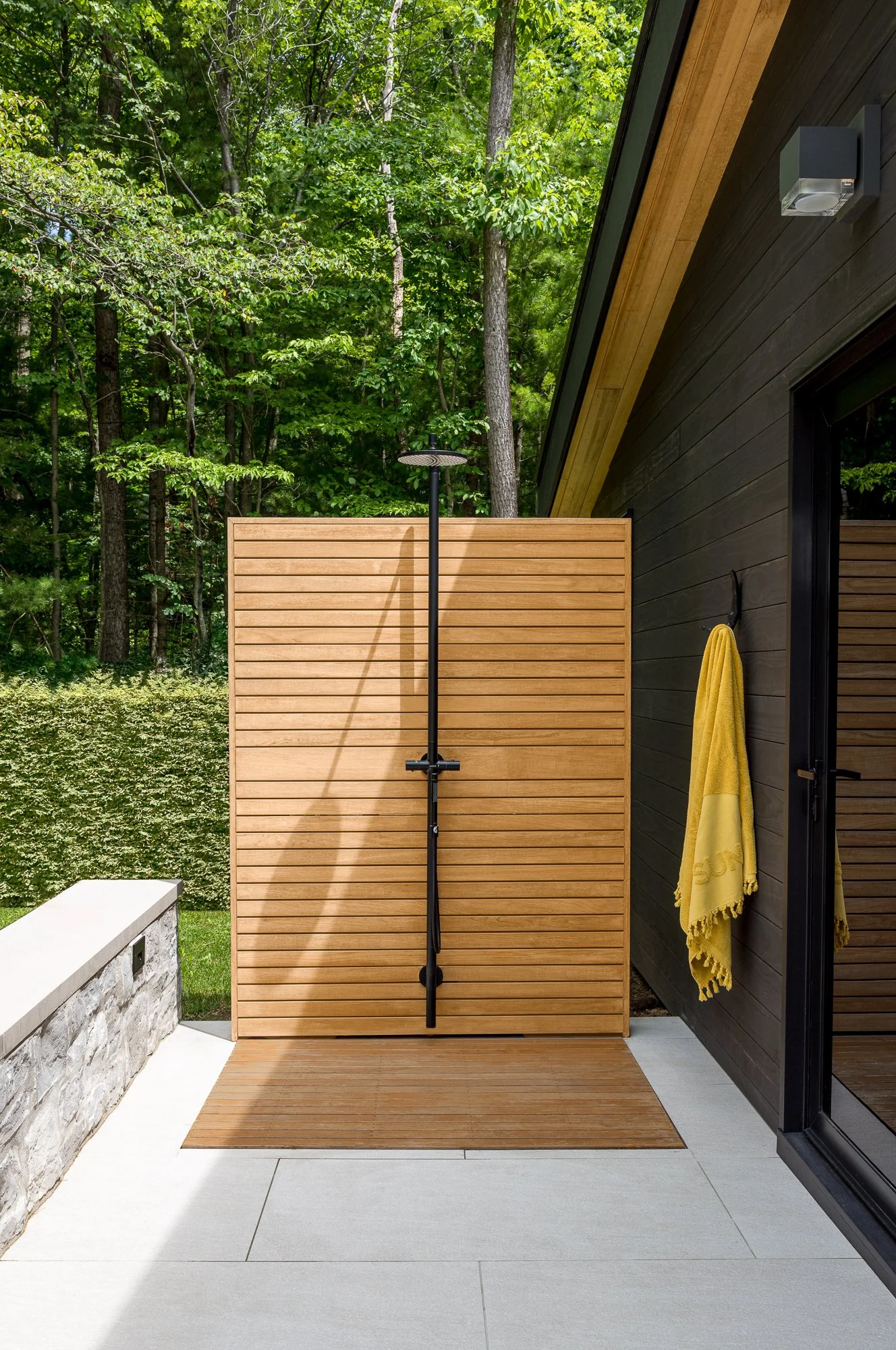 Outdoor shower area with a black shower fixture mounted on a wooden privacy screen, yellow towel hanging on the house wall, surrounded by lush green trees and modern black house exterior.