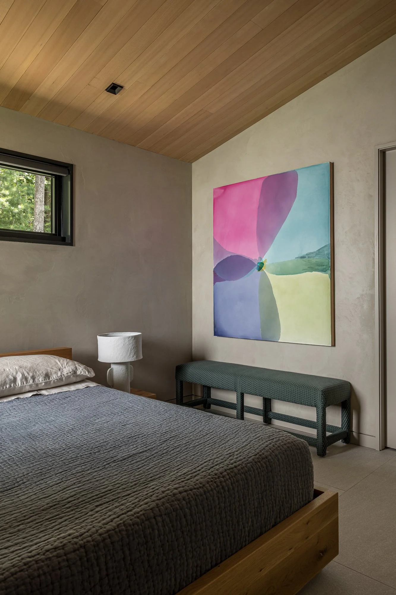A bedroom with a wooden ceiling, a window with trees outside, a bed with gray textured bedding, a white textured lamp, a colorful abstract painting on the wall, a gray bench with a patterned cushion, and sliding closet doors.