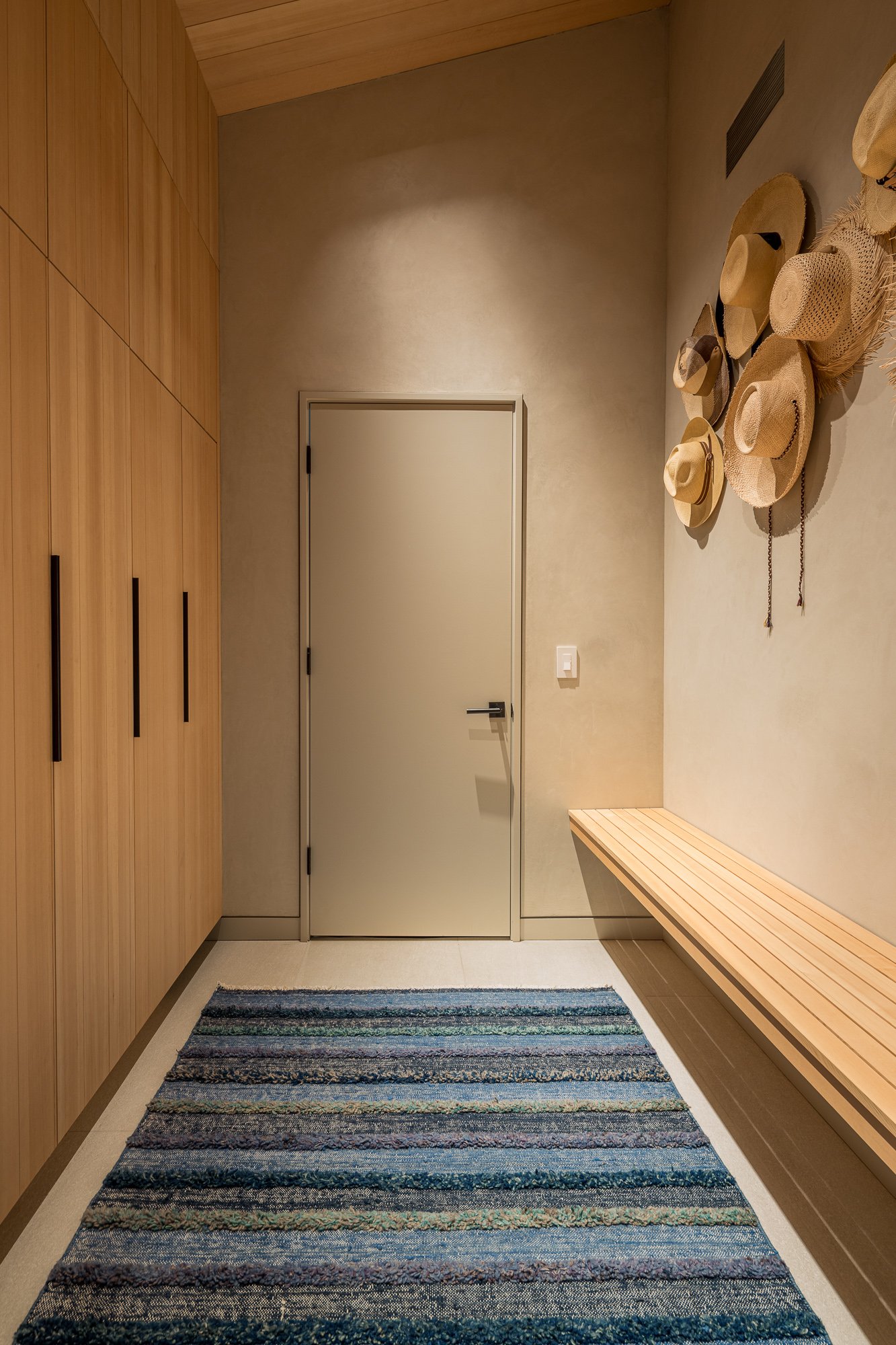 Interior of a room with wooden cabinets on the left, a white door in the center, a bench on the right, a wall with hats hanging, and a striped rug on the floor.