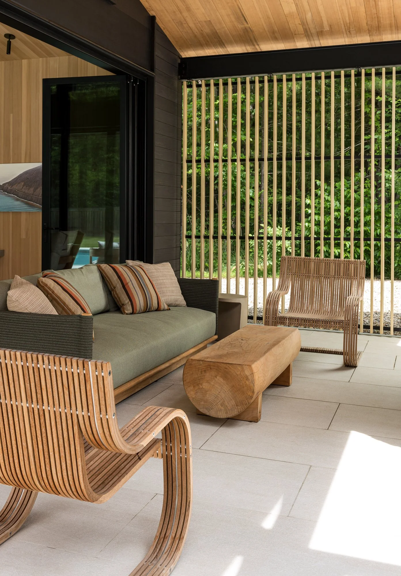 Modern outdoor patio with wooden furniture, including a sofa with cushions and a log-shaped coffee table, surrounded by a wooden slat privacy screen, with a background of lush green trees.