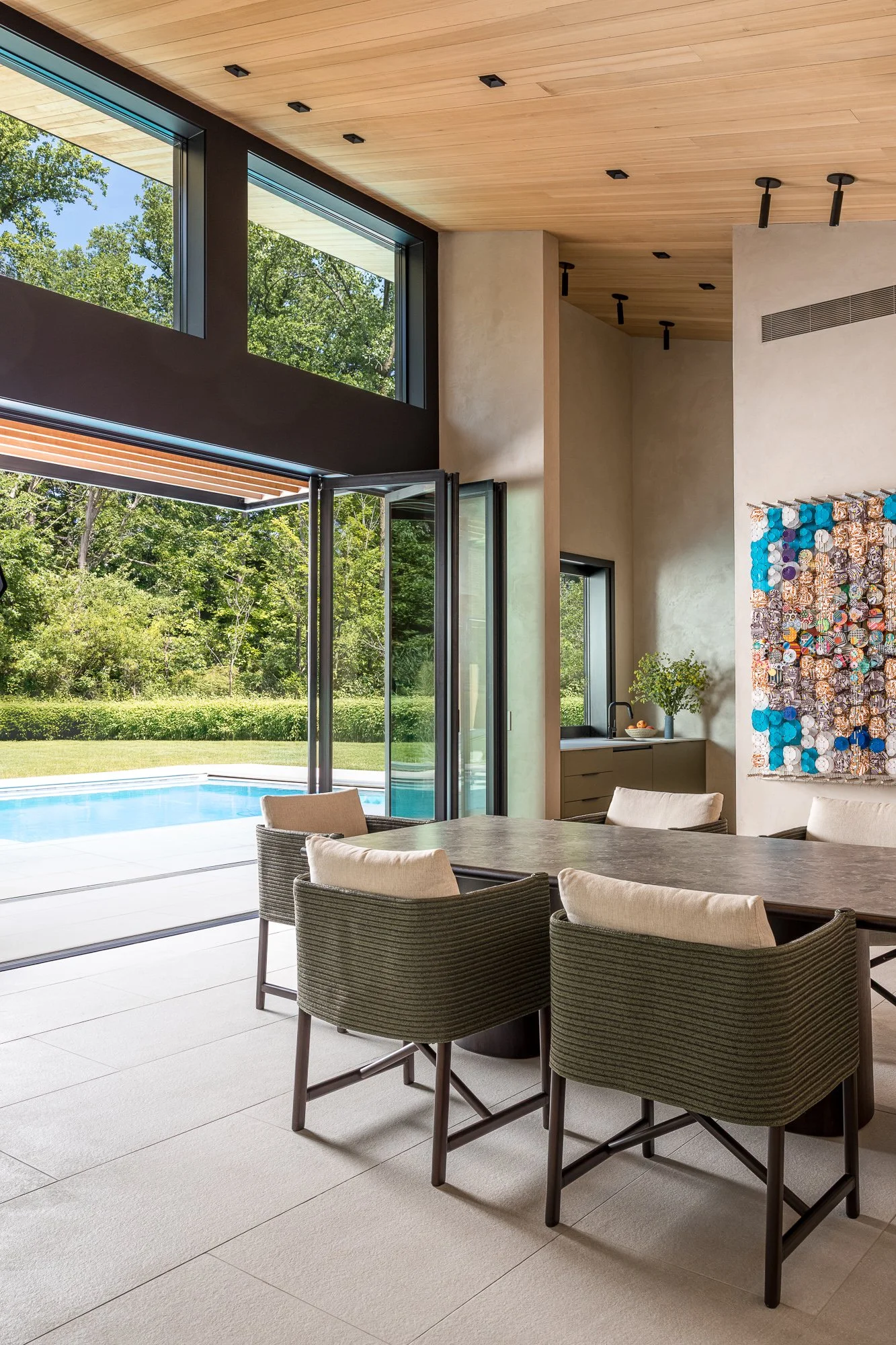 Modern living room with large windows, view of outdoor pool and greenery, indoor seating around a dining table, and contemporary wall art.