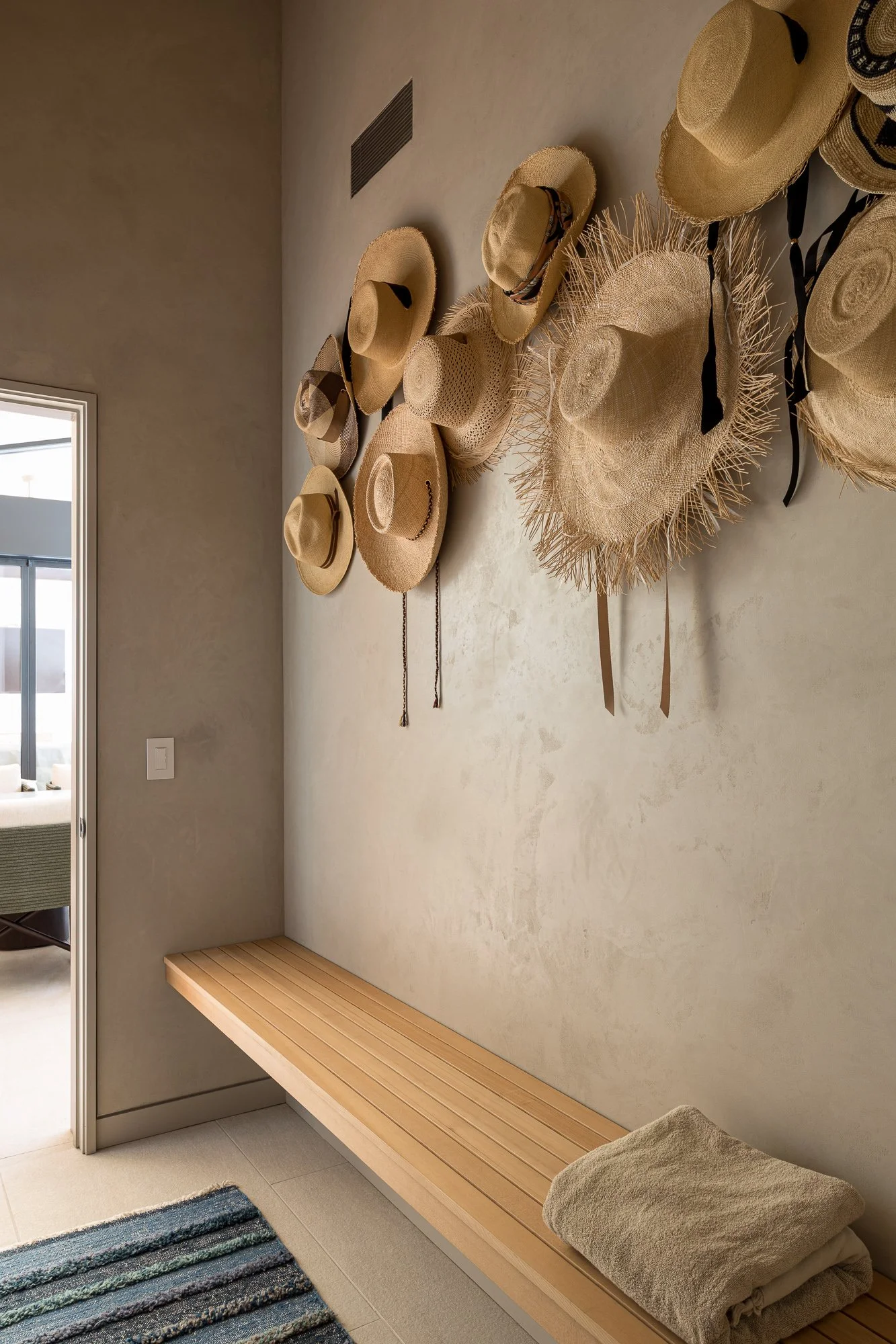 Entryway with a long wooden bench, beige wall decor including multiple woven hats, some with black ribbons, and a rolled-up beige towel on the bench. A colorful striped rug partially visible in the foreground, and a doorway leading to a living space 