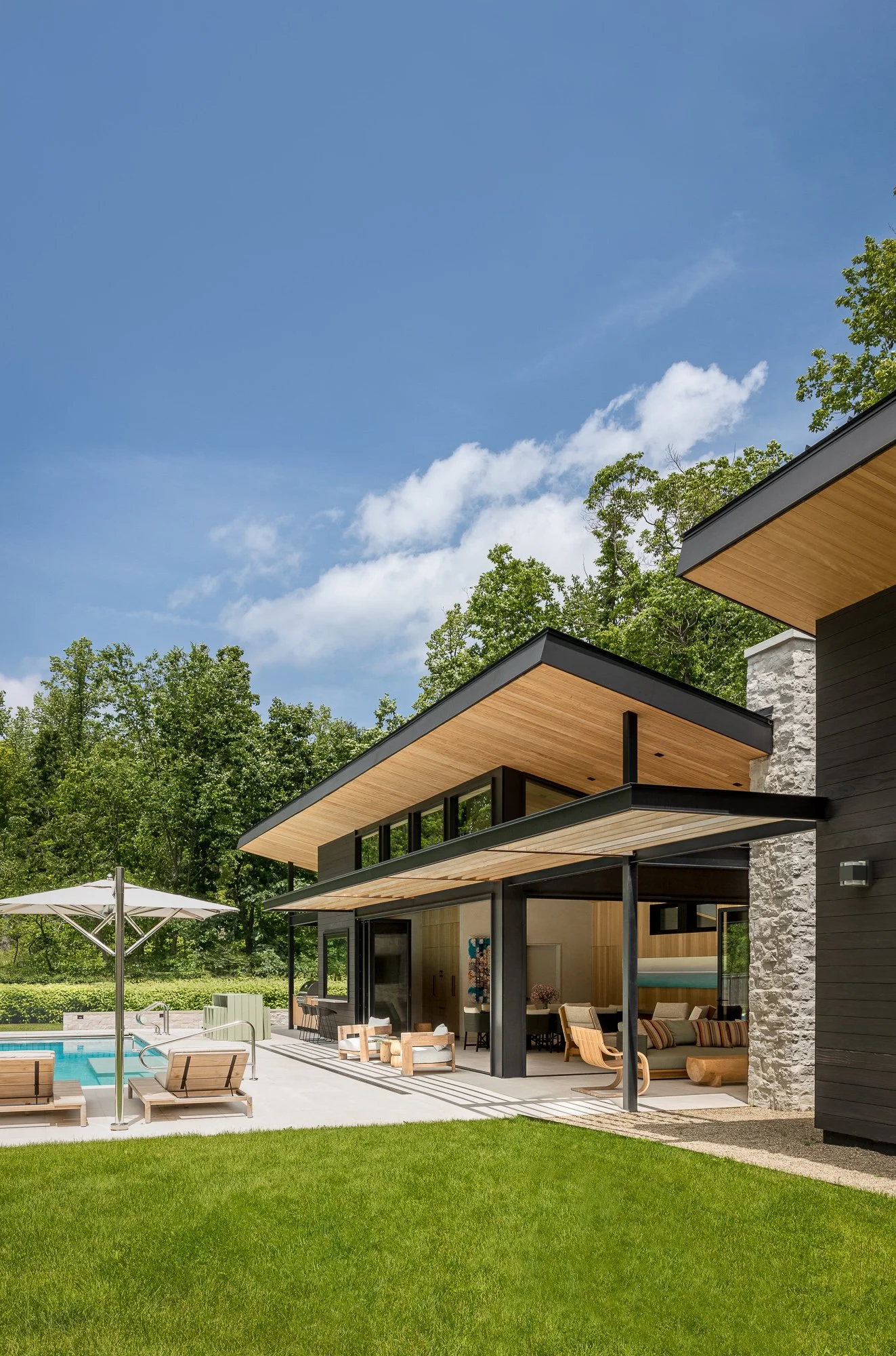 Modern house with large windows, wooden accents, outdoor patio, poolside chairs, and patio umbrella set in a lush green yard under a bright blue sky.