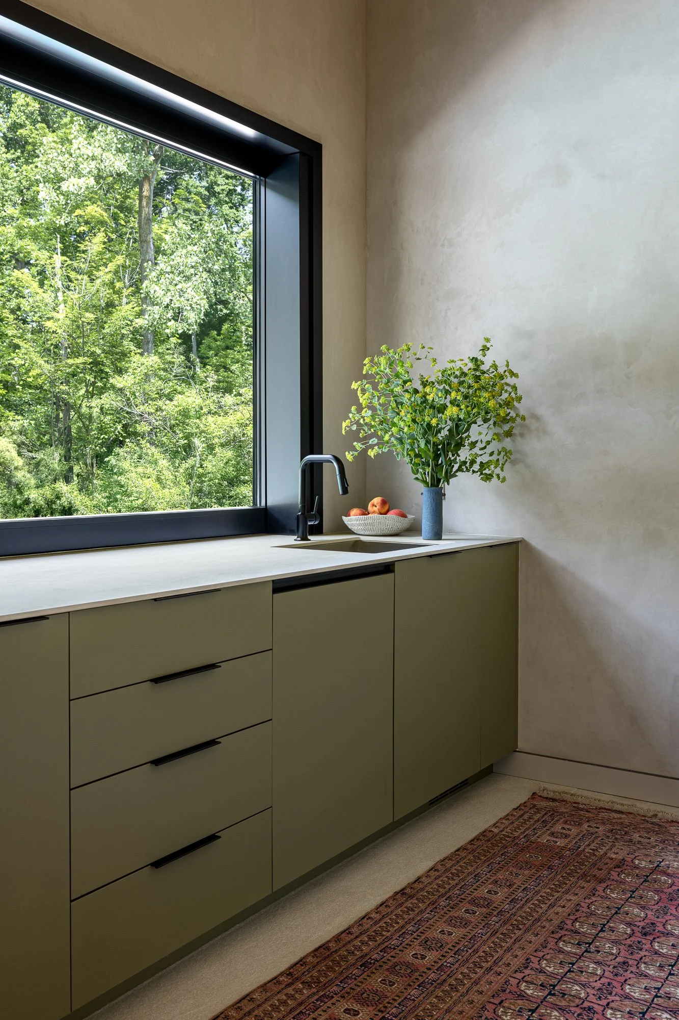 A kitchen with a large window overlooking green trees, a beige countertop, a black faucet, a vase with green plants, a bowl of peaches, a patterned rug, and beige cabinets.