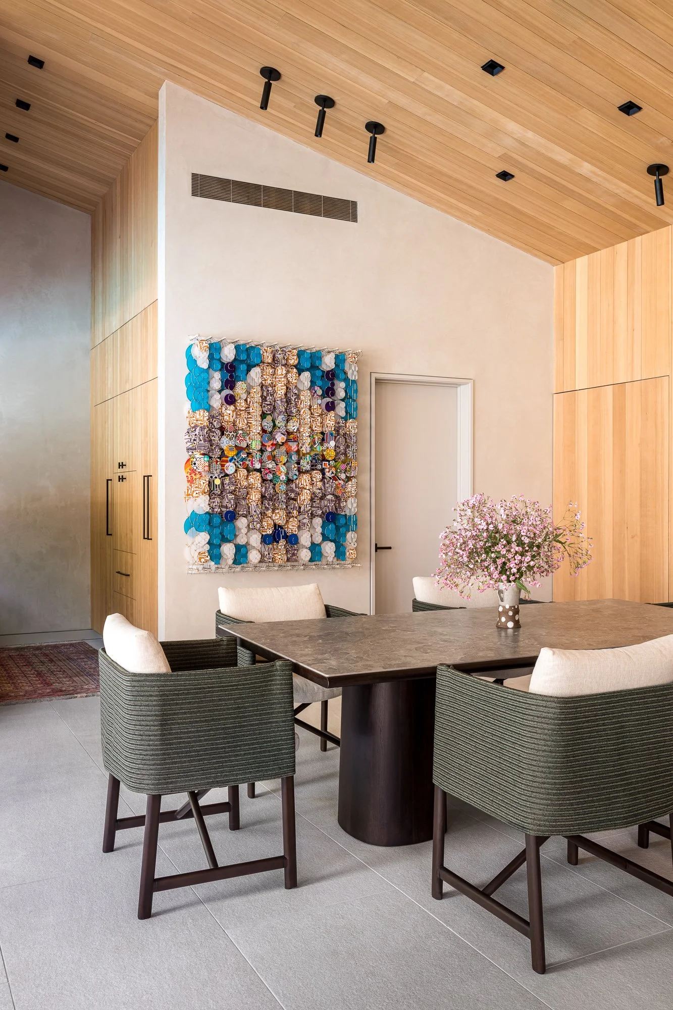 Interior of a modern dining room with a wooden ceiling and wall panels, a painting made of bottle caps hanging on the wall, a dining table with a flower vase, and four upholstered chairs.