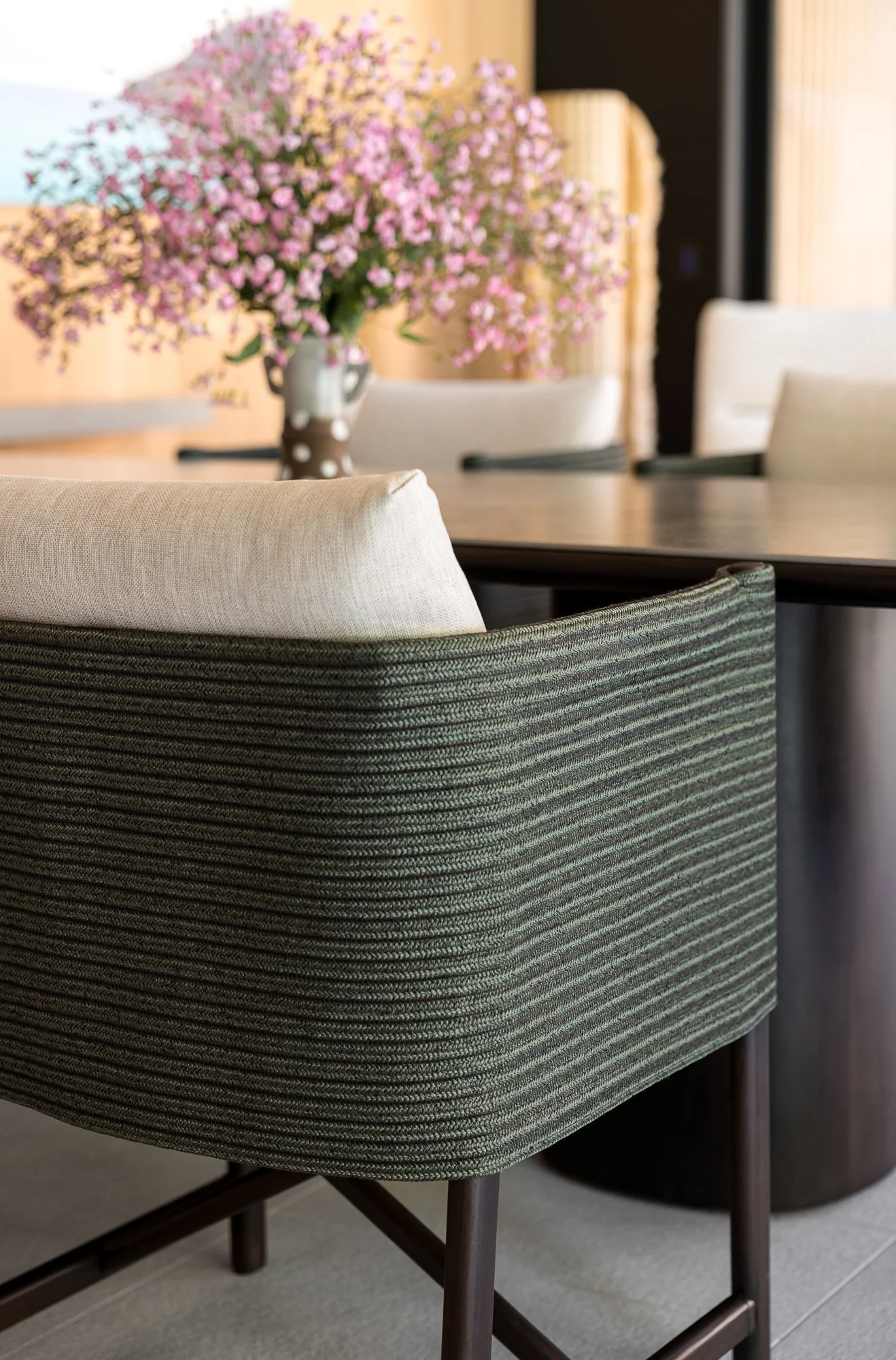 Close-up of a modern dining chair with dark wooden legs and a fabric seat and backrest, with a blurred background of a dining room table and a vase with pink flowers.