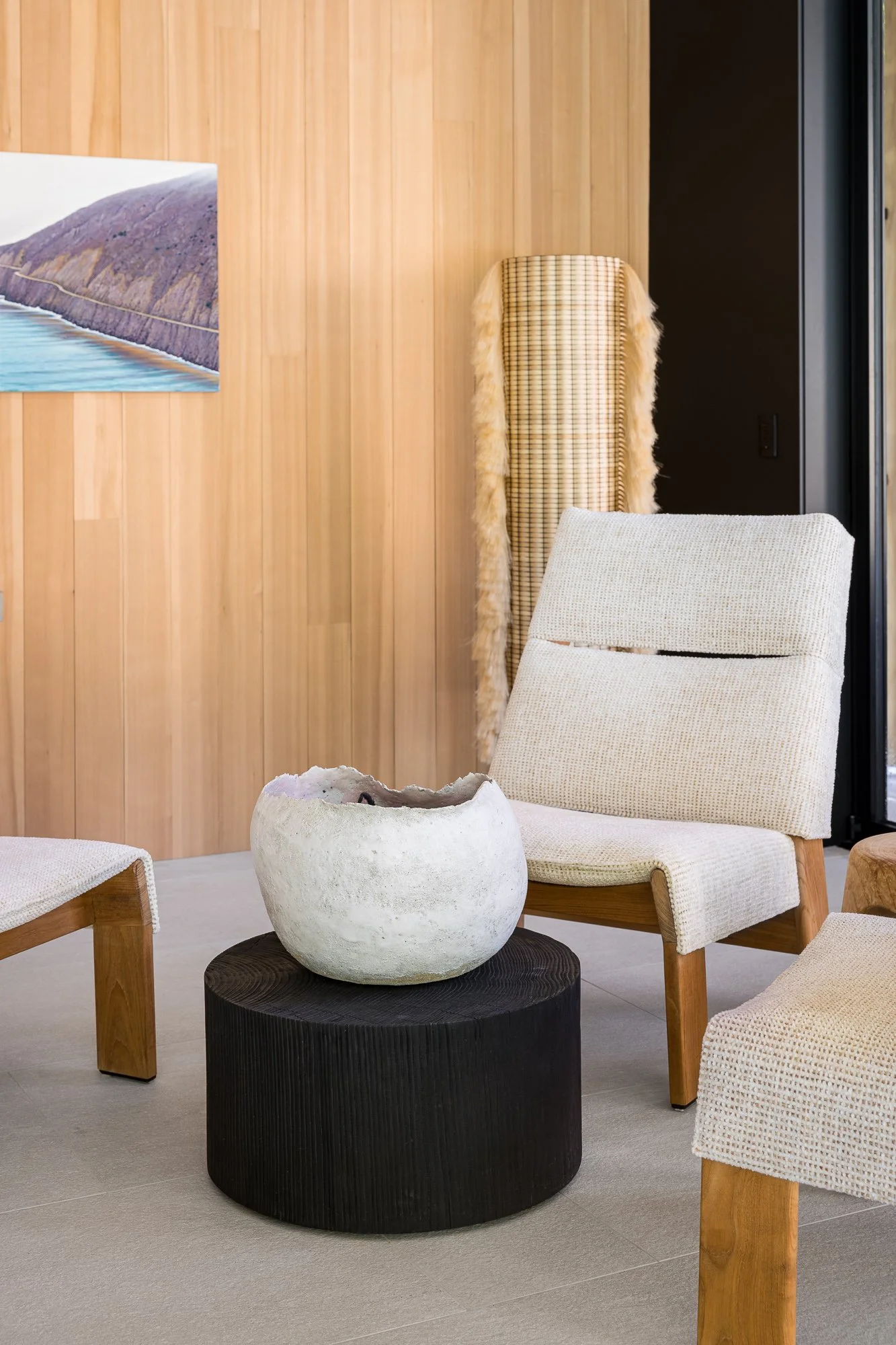 Modern living room with light wood panel walls, cream-colored armchair, black round table with white ceramic bowl, wooden chairs, beige carpet, and a partial wall with artwork.