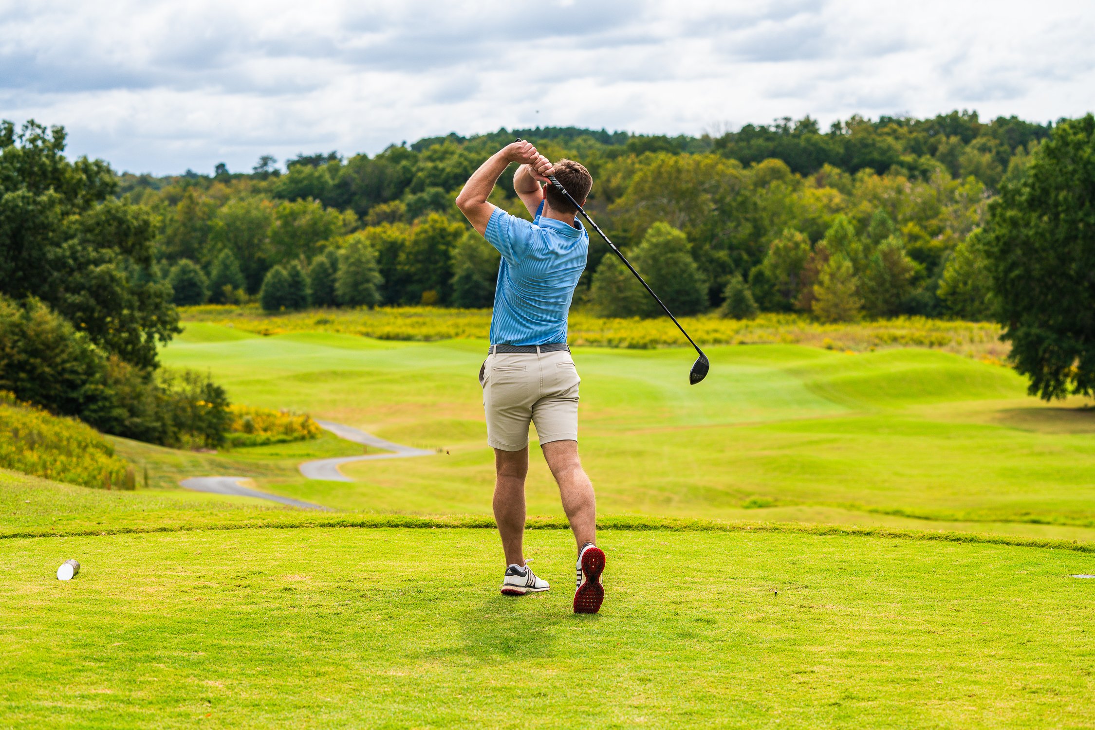 Golfer driving a shot off the tee, facing a green fairway with winding cart paths and distant hills.