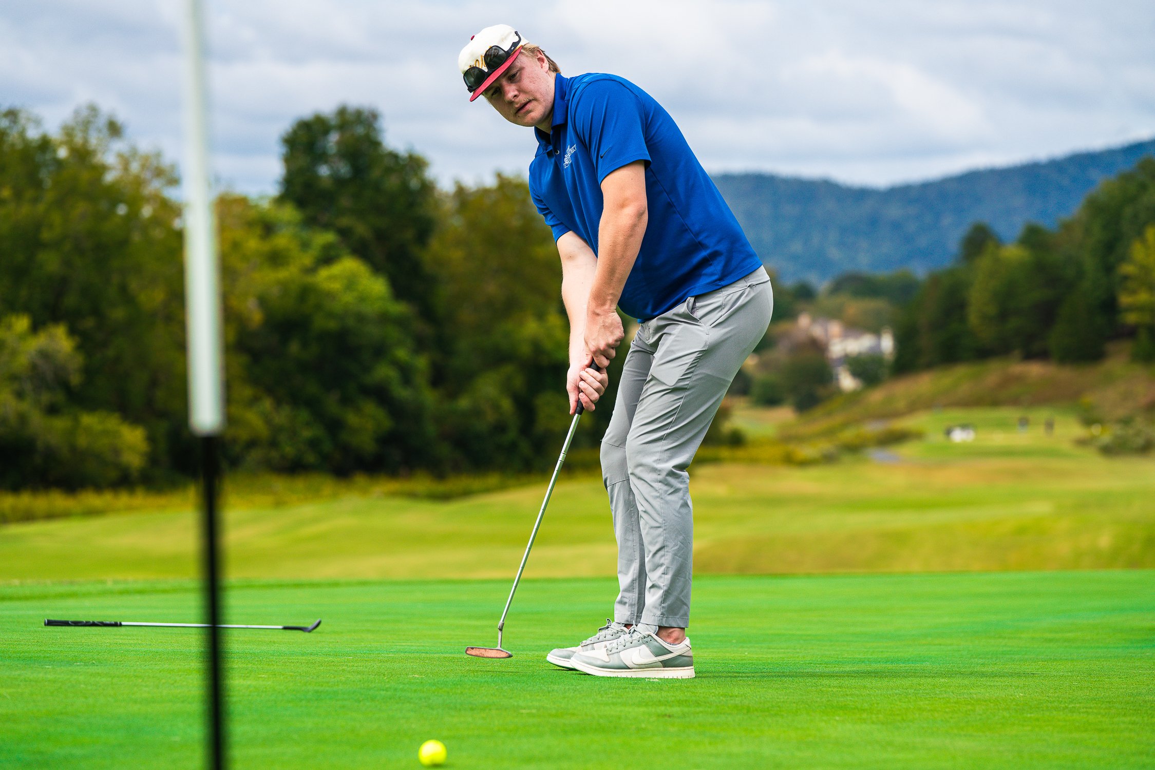 Golfer preparing to putt while two others observe, standing on a well-maintained green with trees in the background.