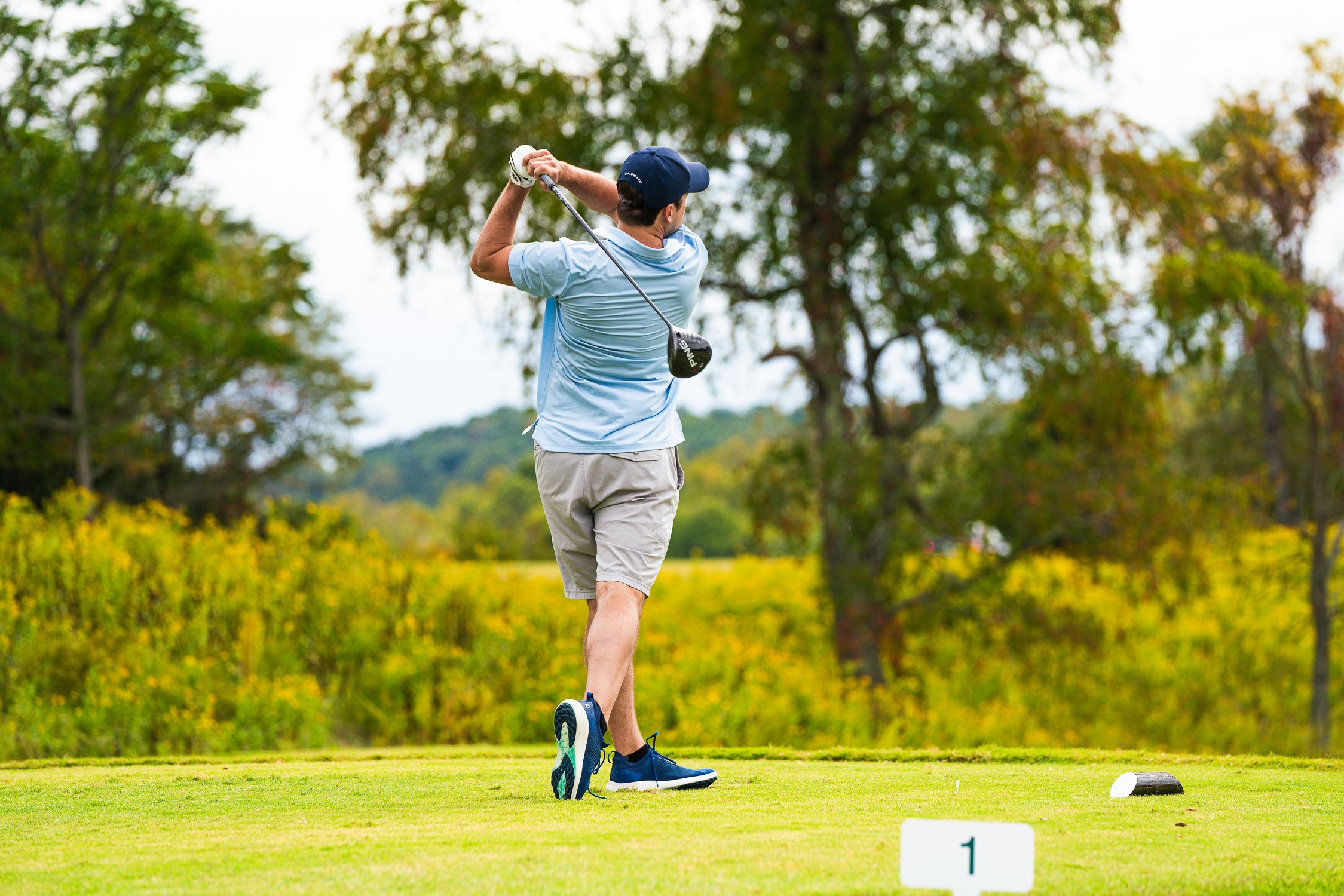 Golfer driving a shot off the tee, facing a green fairway with winding cart paths and distant hills.