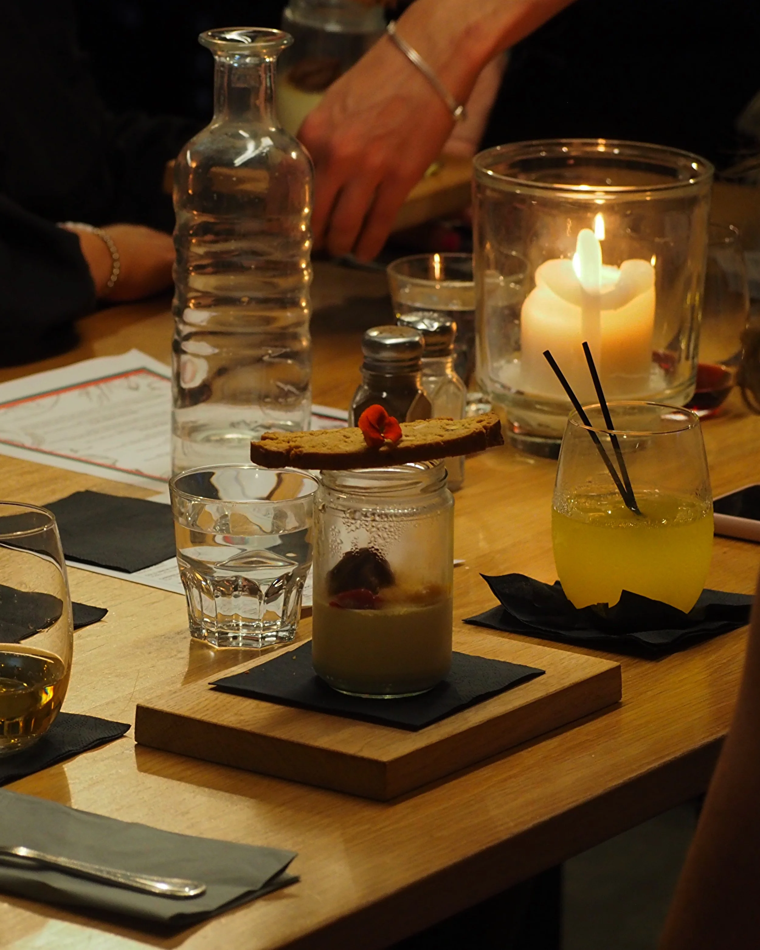Candlelit table with dessert jar, biscotti, cocktails, and water bottle.