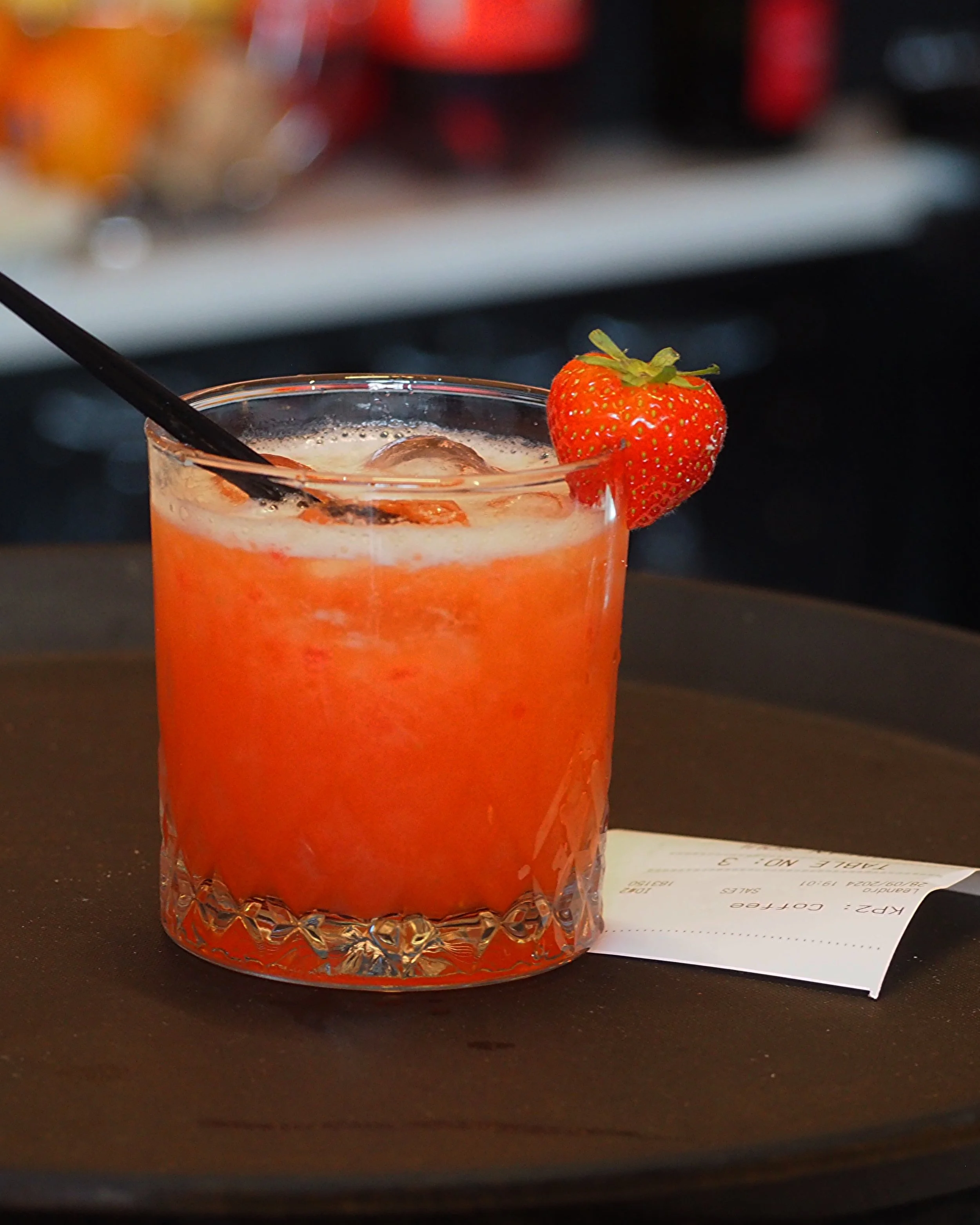 A glass of orange-hued cocktail with ice, garnished with a strawberry and a black straw, placed on a tray with a receipt.