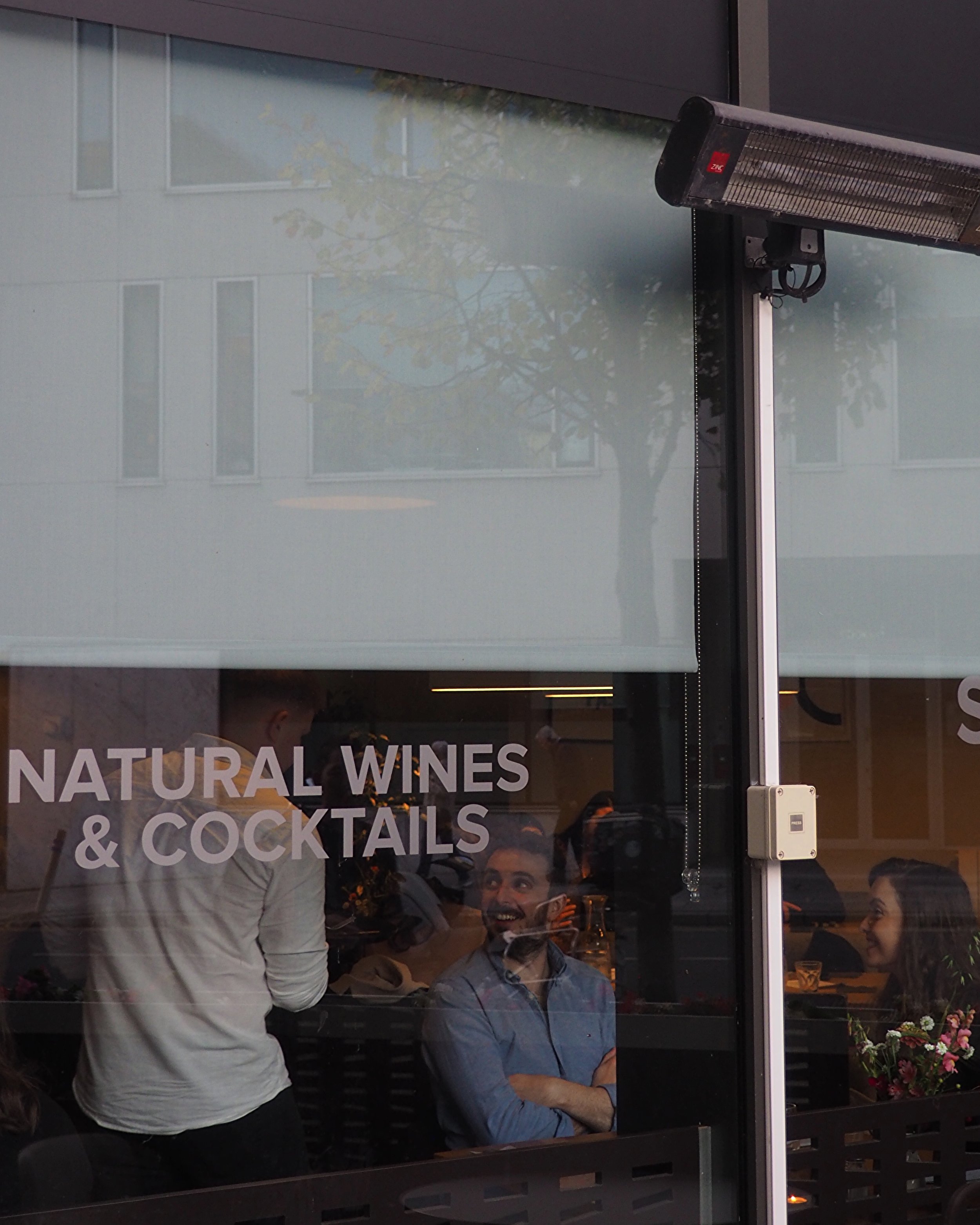 People in a cafe with sign reading 'Natural Wines & Cocktails' on the window, chatting and smiling.