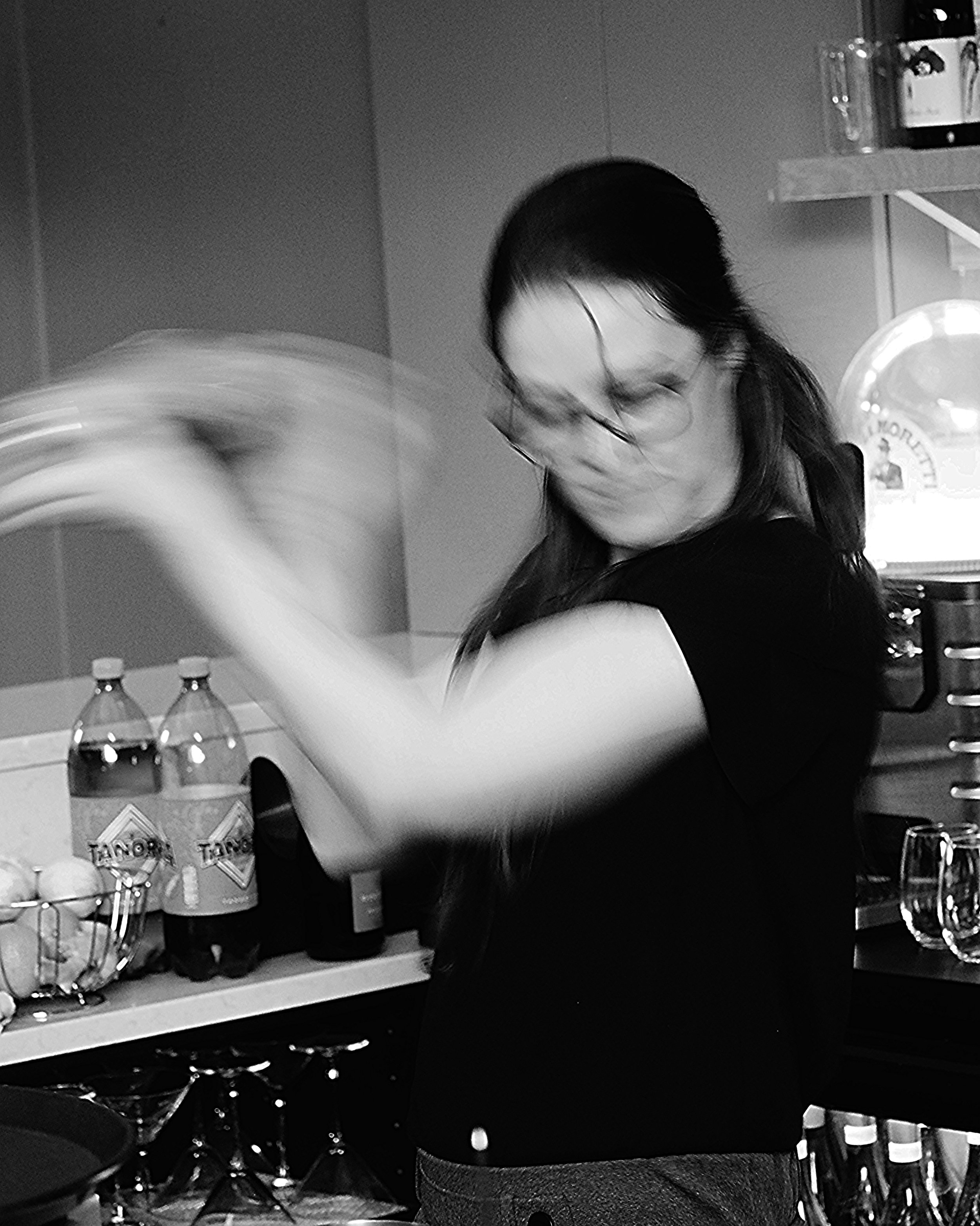 A blurry image of a woman shaking a cocktail shaker, with bottles and glasses in the background.