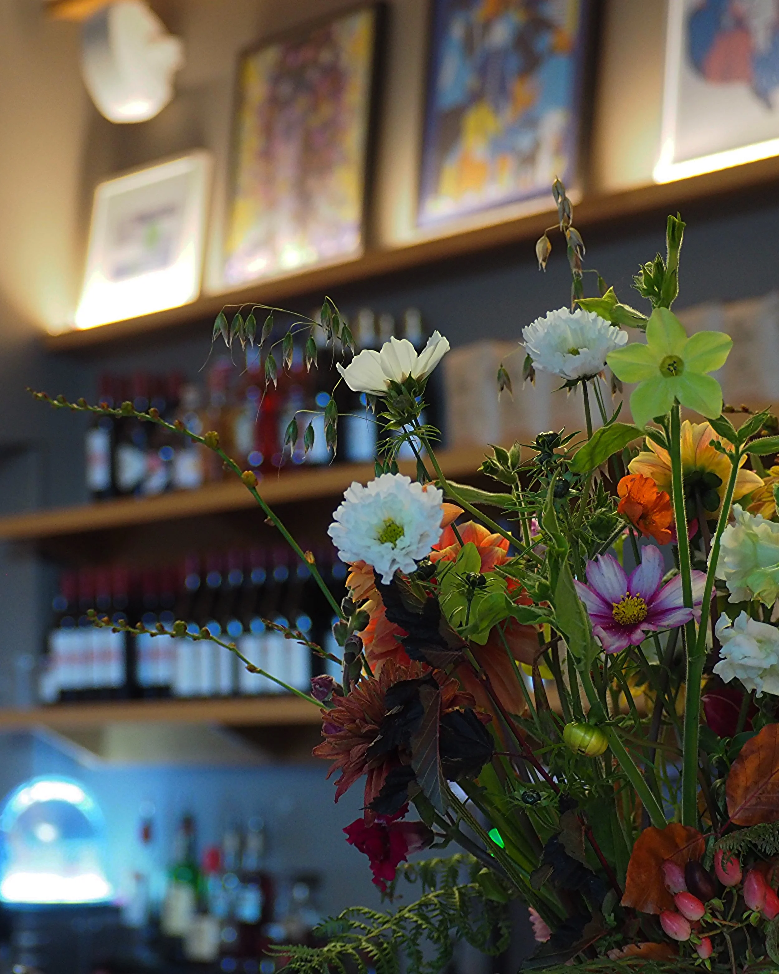 Colorful flower arrangement in a bar setting with shelves of bottles and framed artwork on the wall.