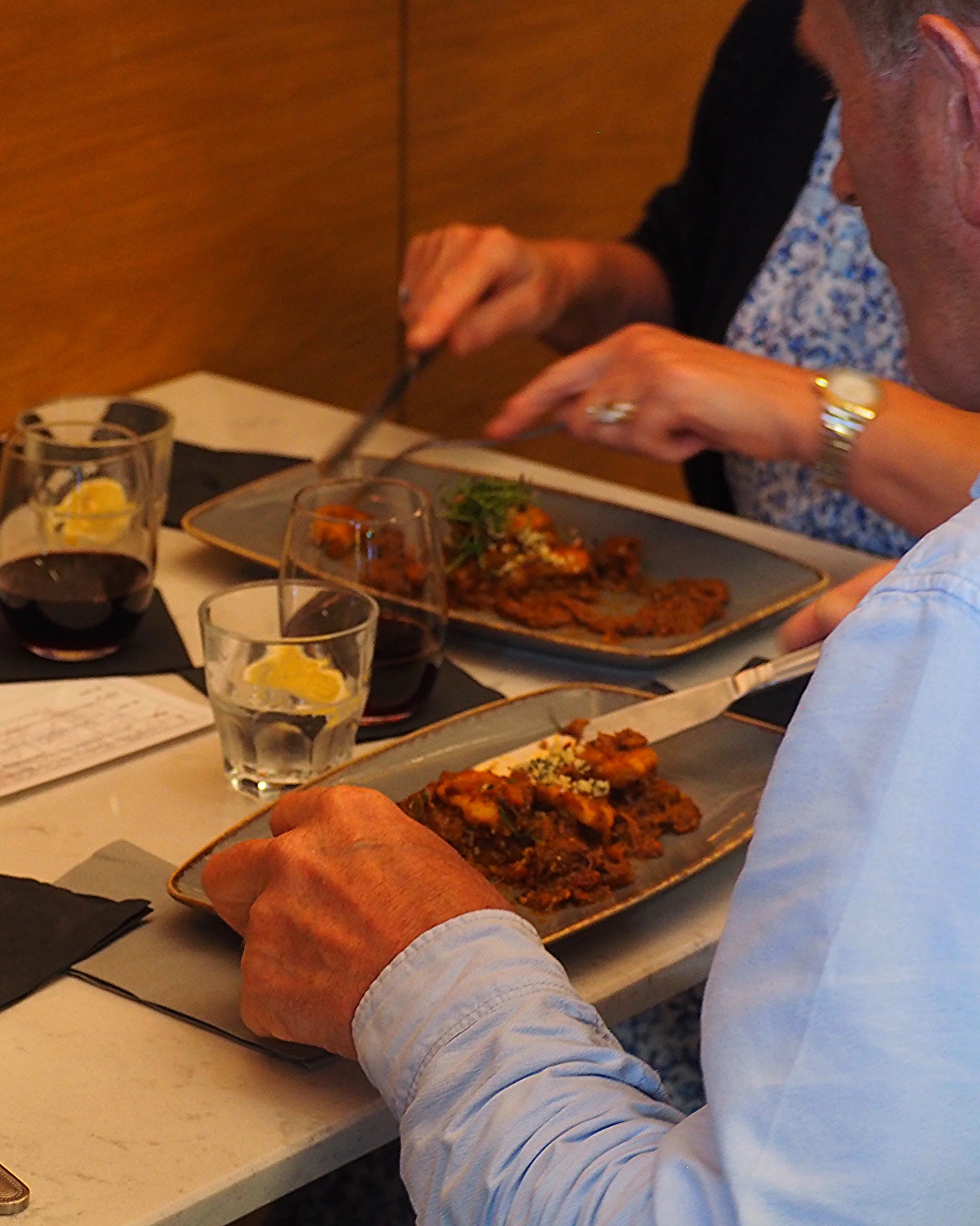 Two people dining at a restaurant table, enjoying plated meals with glasses of red wine and water with lemon. A notepad is visible on the table.