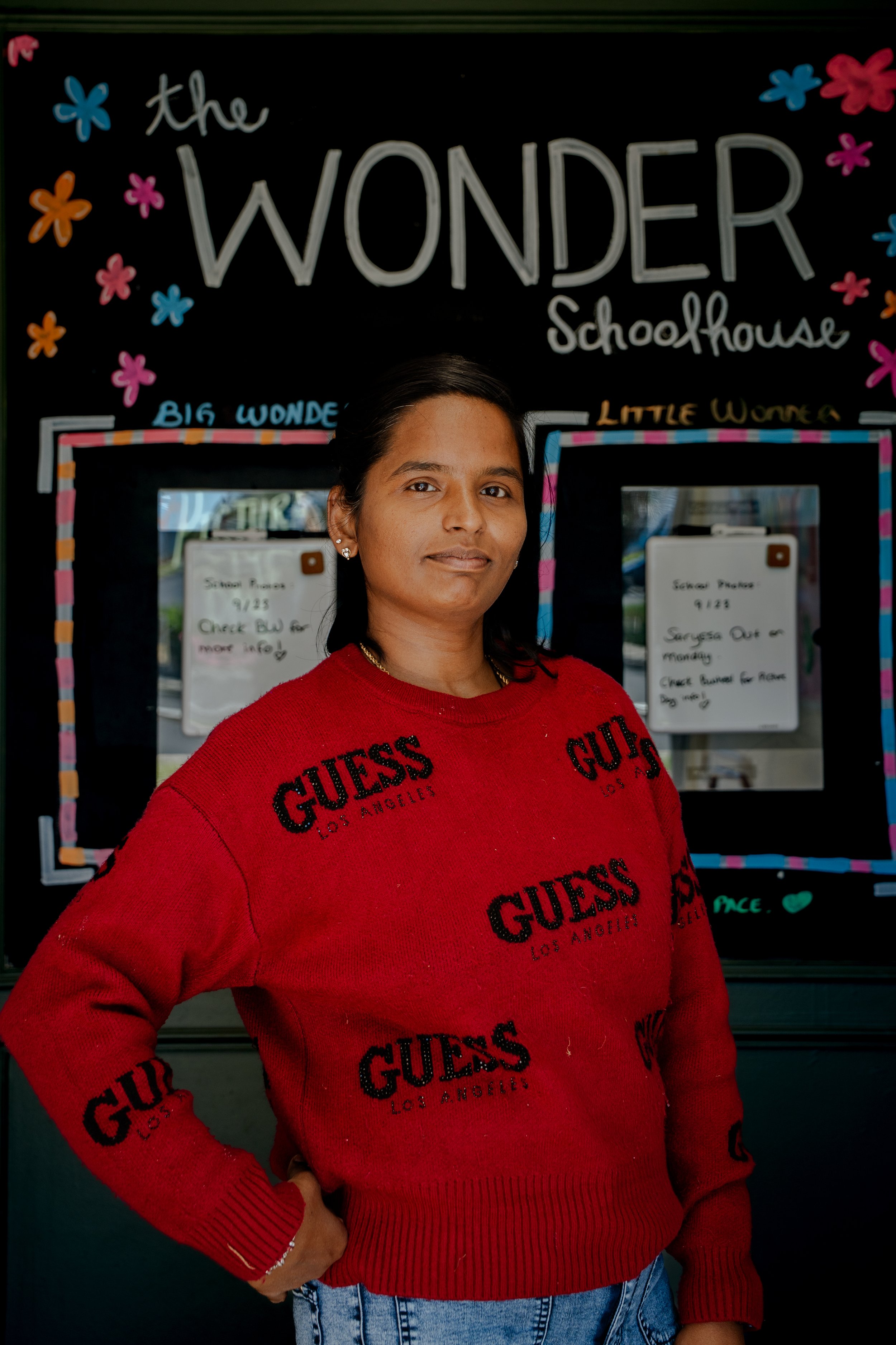 A woman standing in front of a blackboard with colorful drawings and text that says 'the WONDER Schoolhouse'. She is wearing a red GUESS sweatshirt and has dark hair tied back.