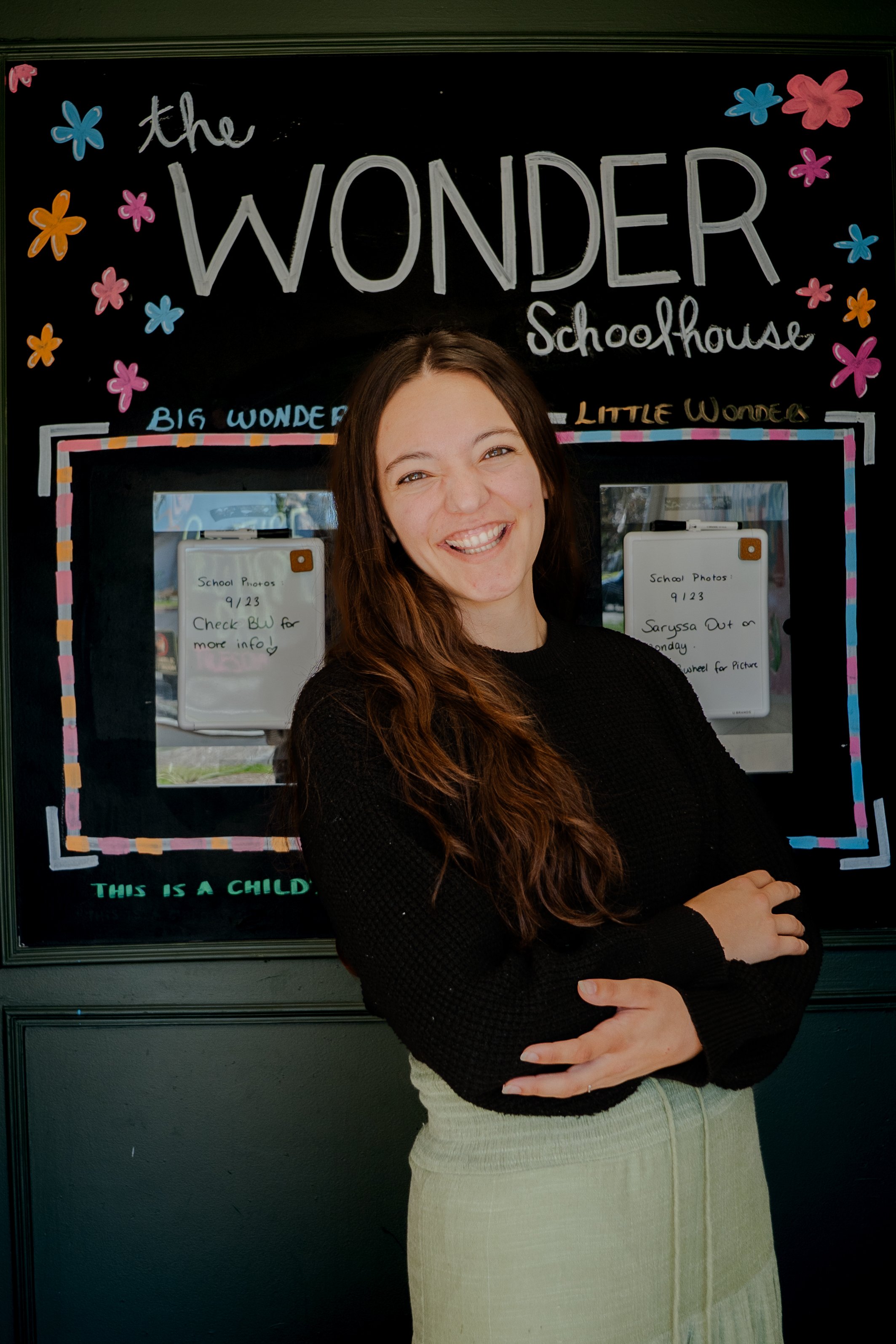 A young woman with long wavy brown hair smiling and crossing her arms in front of a blackboard with colorful decorations and text reading 'the wonder schoolhouse'.