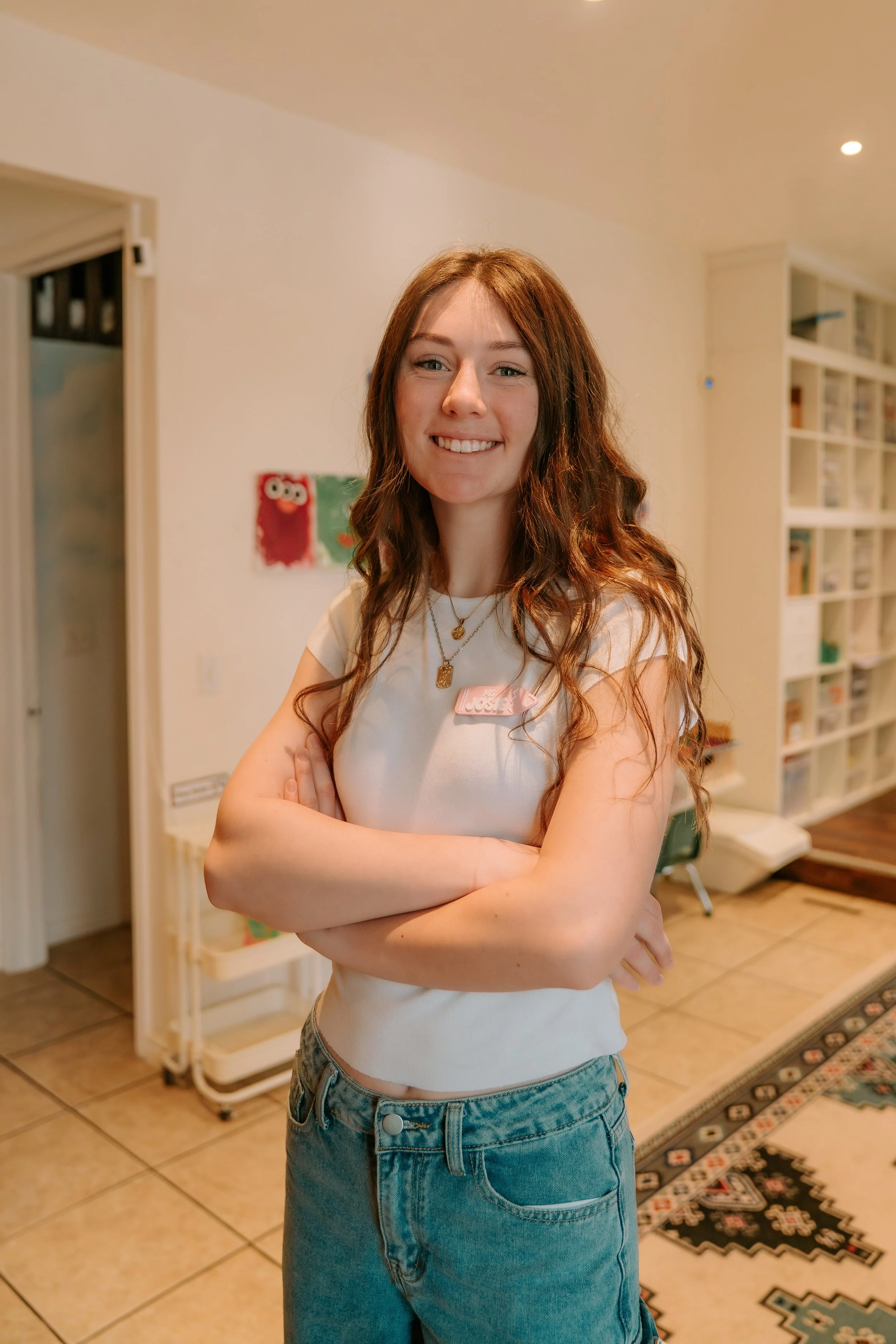 A young woman with long, wavy auburn hair smiling and crossing her arms, standing in a cozy room with a colorful chalkboard, bookshelves, and a patterned rug.