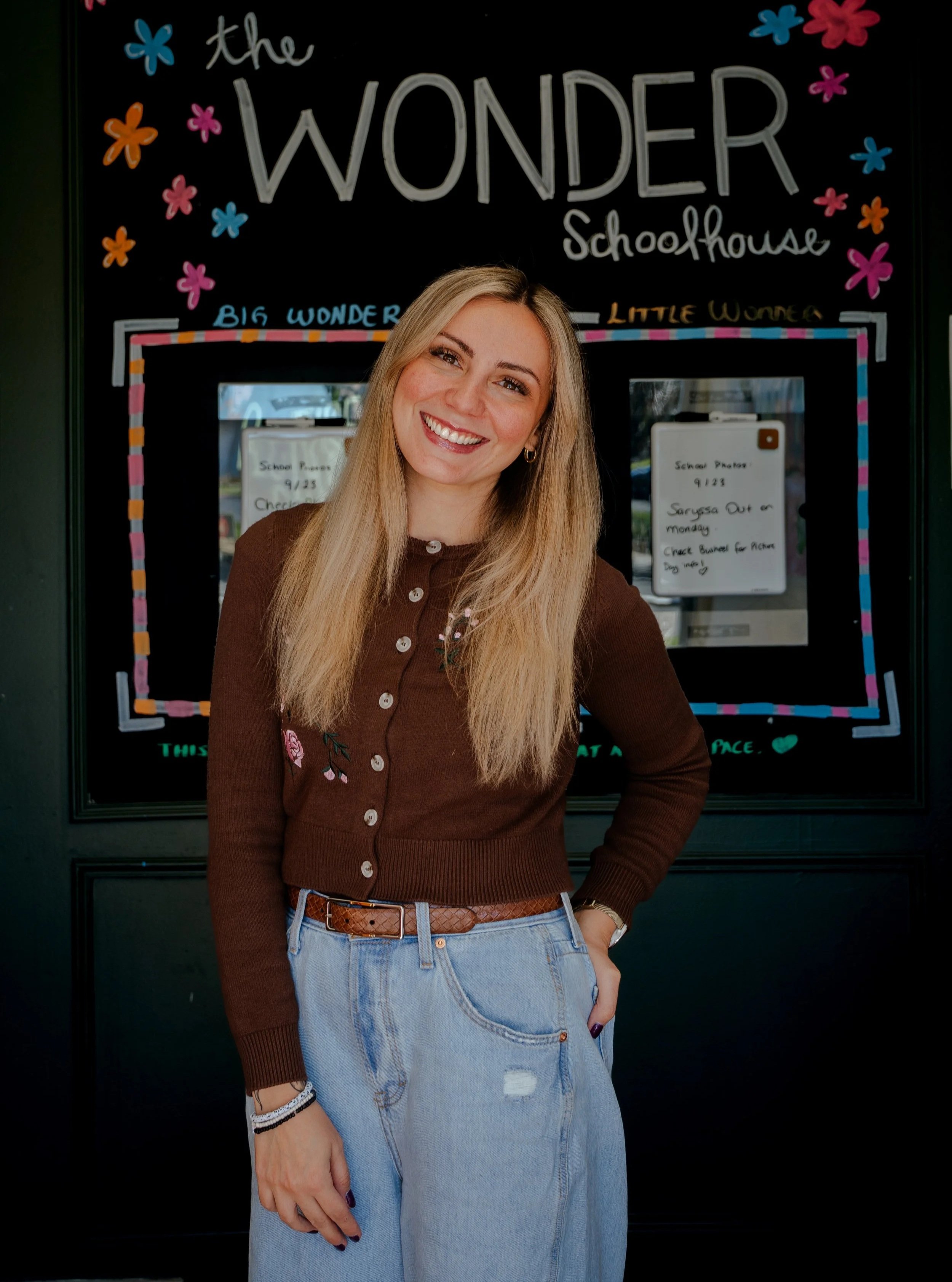 A smiling young woman with long blonde hair wearing a brown cardigan and light blue jeans standing in front of a colorful blackboard with the words 'the wonder schoolhouse' written on it.