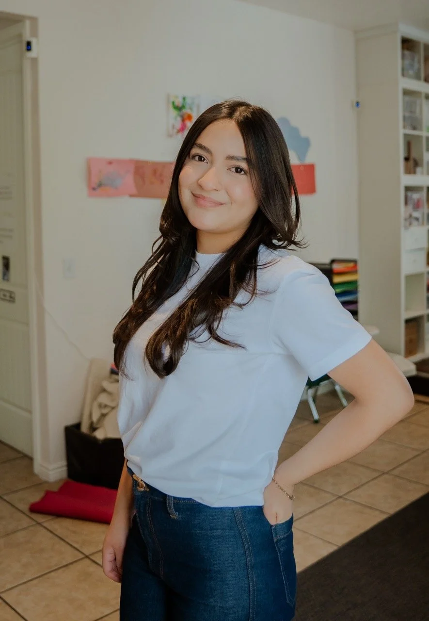 A young woman with long dark hair, wearing a white t-shirt and dark jeans, standing indoors with a gentle smile and hand on her hip.