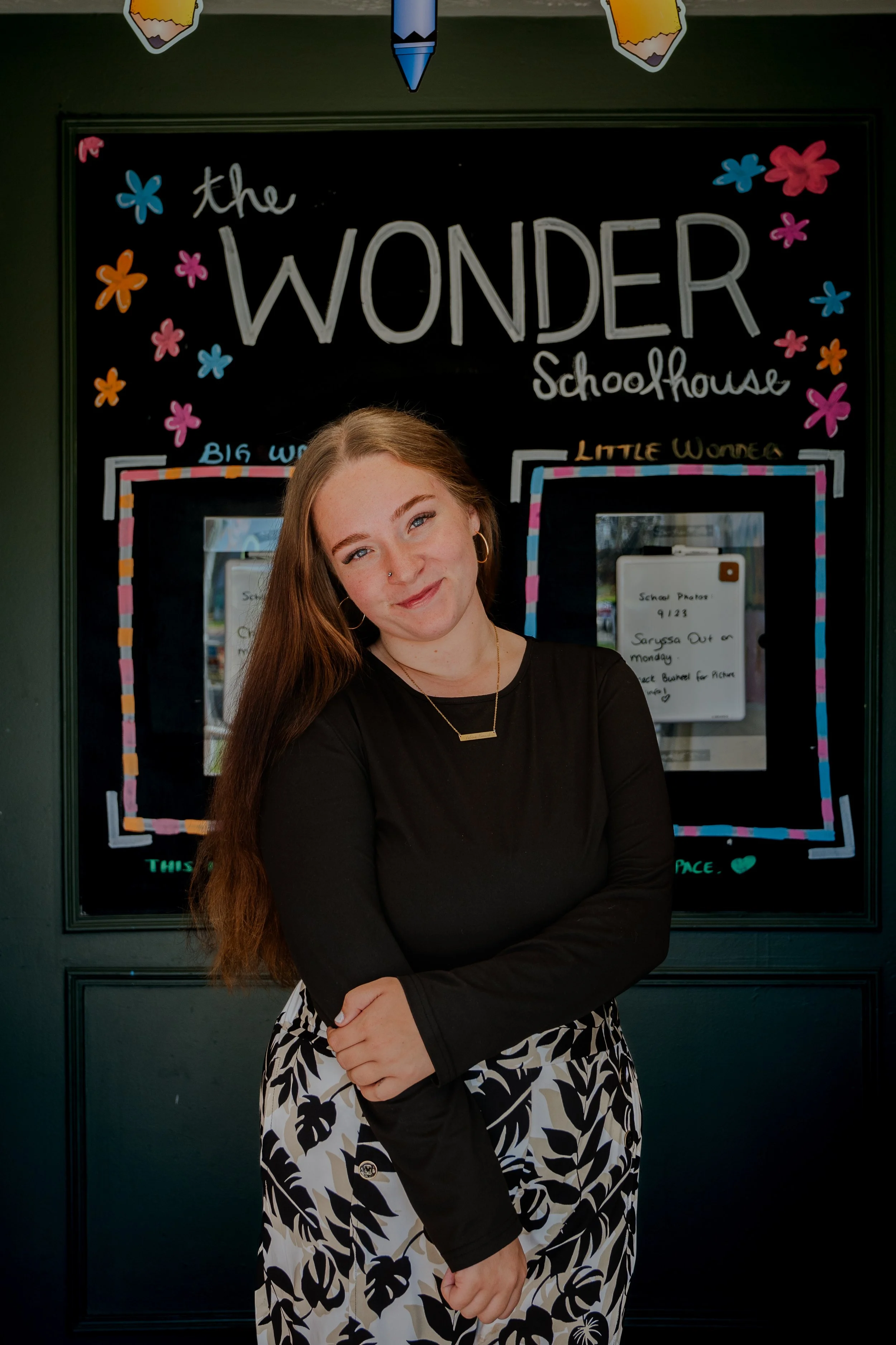 A young woman with long red hair smiling at the camera stands in front of a blackboard with colorful decorations and handwritten text about a schoolhouse theme.