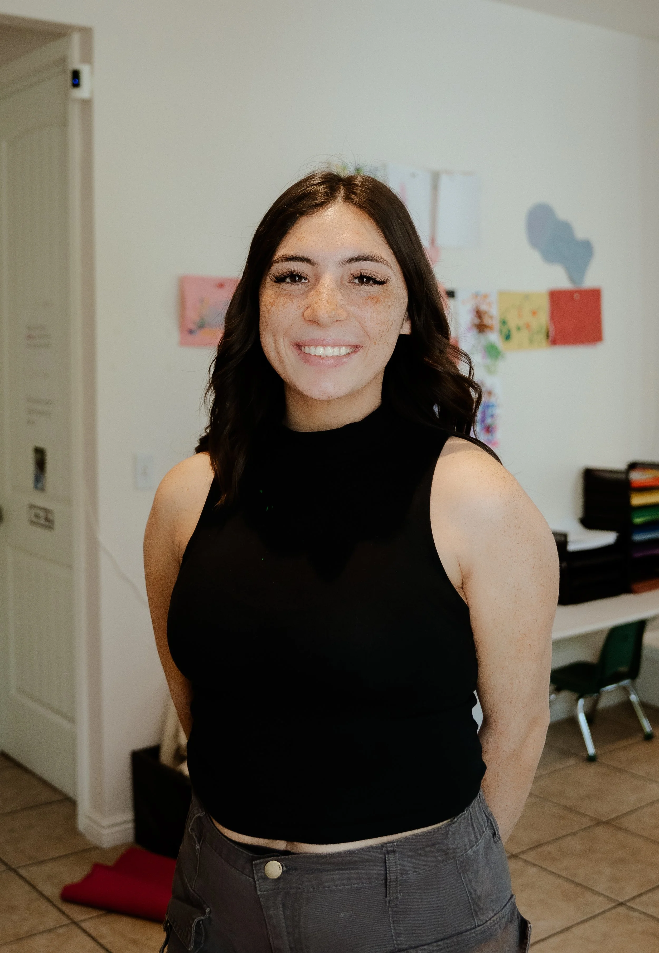 A young woman with dark brown hair and freckles, smiling at the camera in an indoor setting, wearing a black sleeveless top and gray pants.